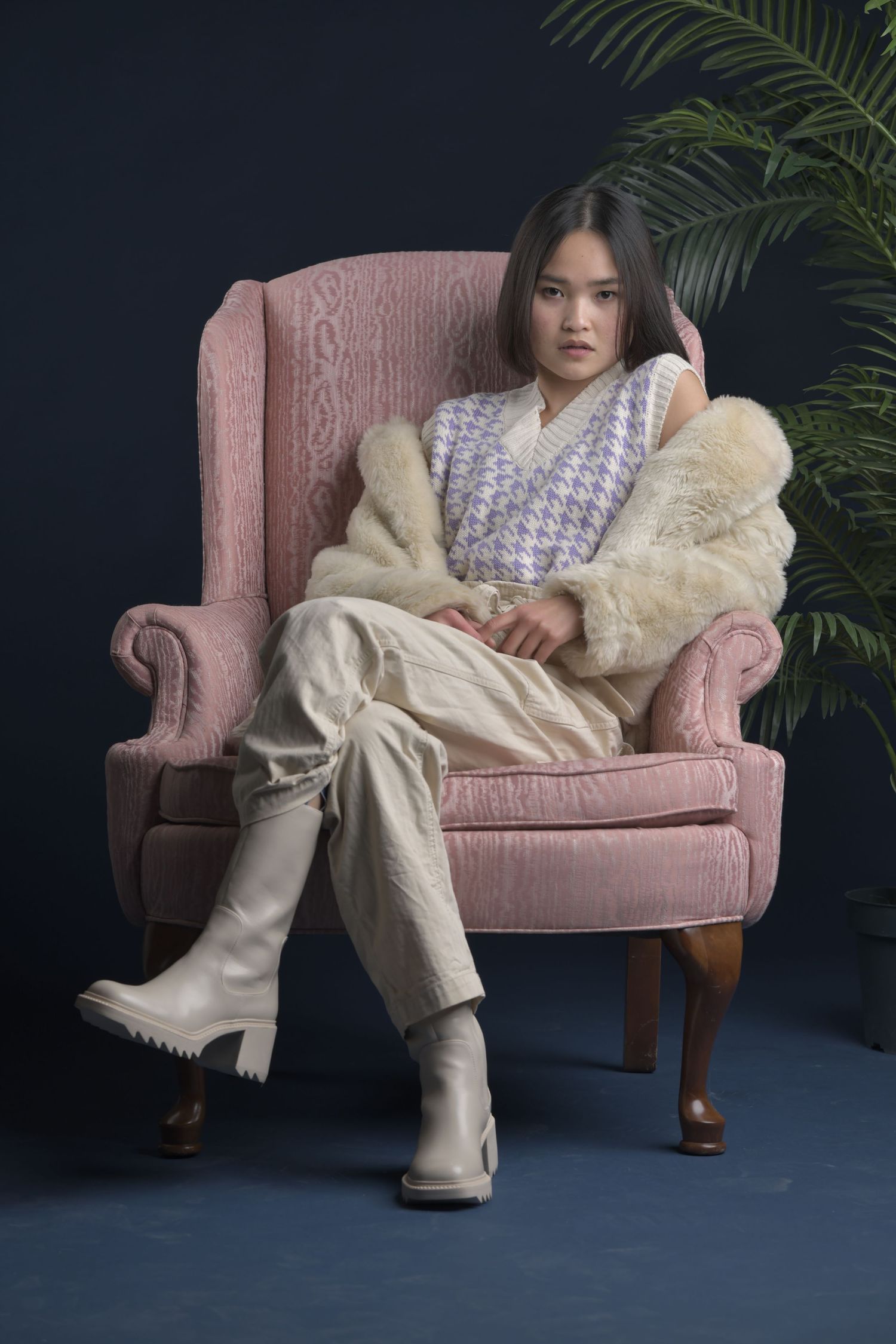 A person in a patterned top and cream pants lounges elegantly across a vintage pink velvet armchair against a dark backdrop.