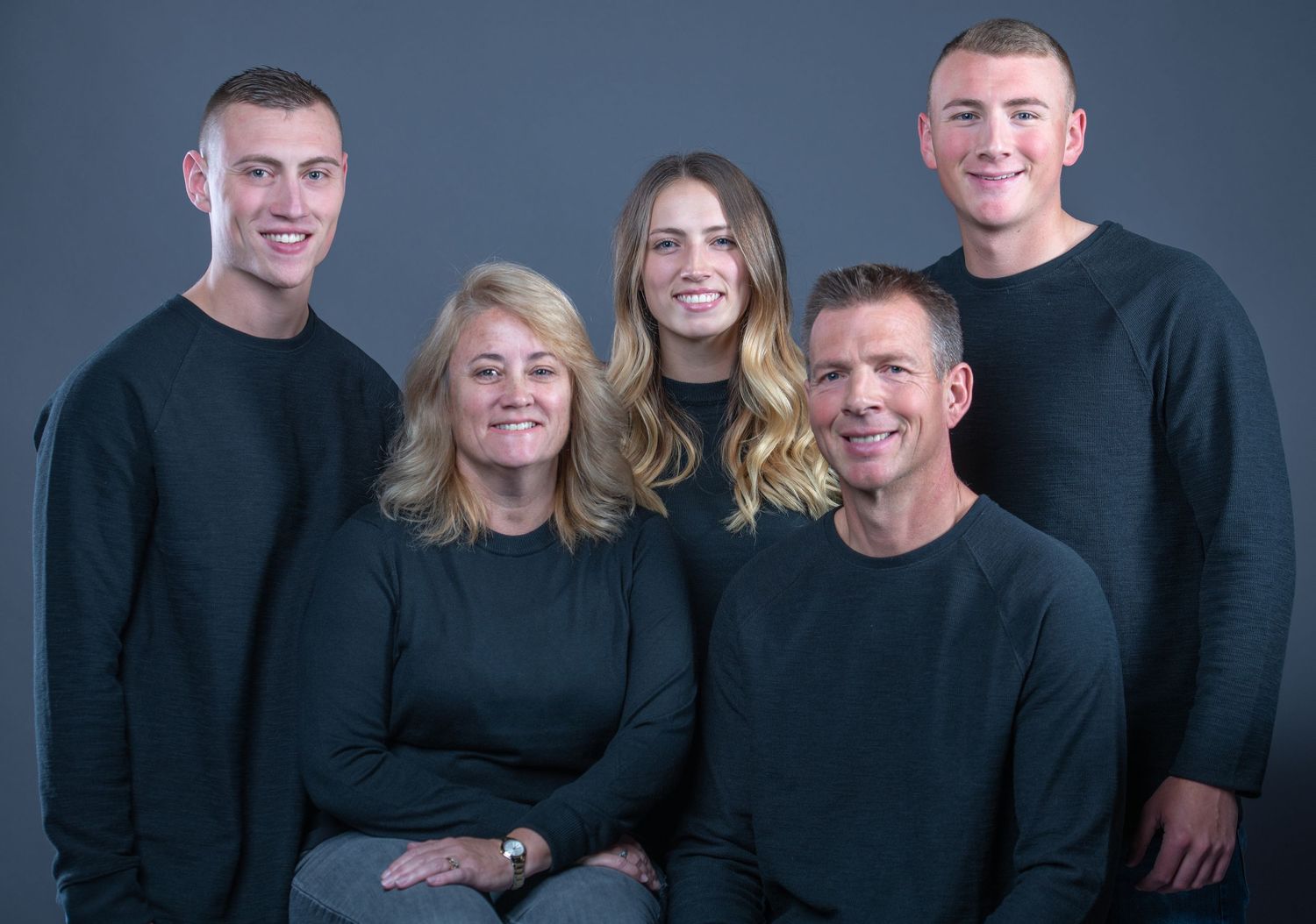 A family group of five people wearing matching black sweaters poses together against a blue-gray background.