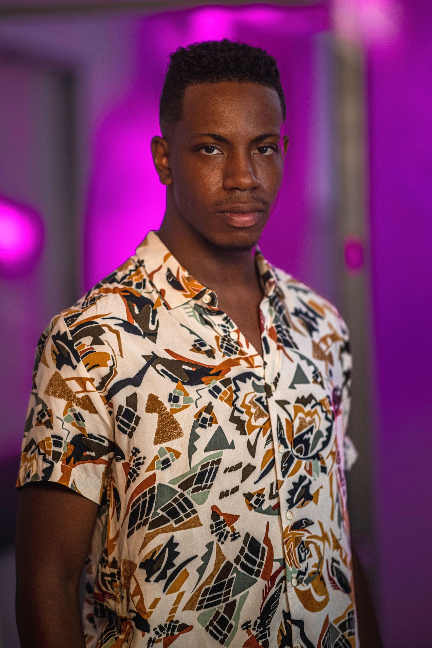 A person wearing a white and brown floral print short-sleeve shirt poses against a purple-lit background.