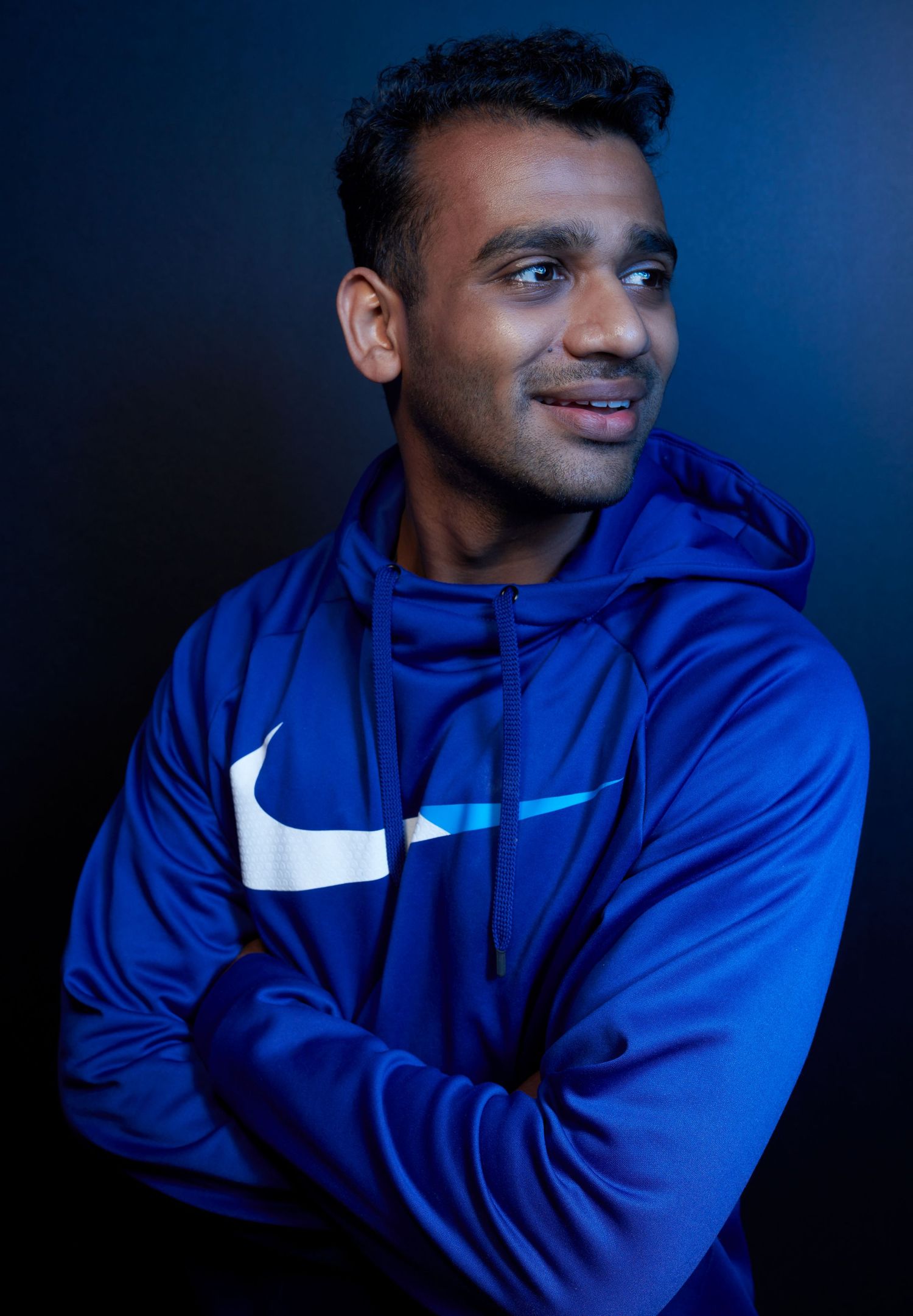 A person wearing a bright blue Nike hoodie smiles warmly against a dark blue backdrop in a professional portrait.