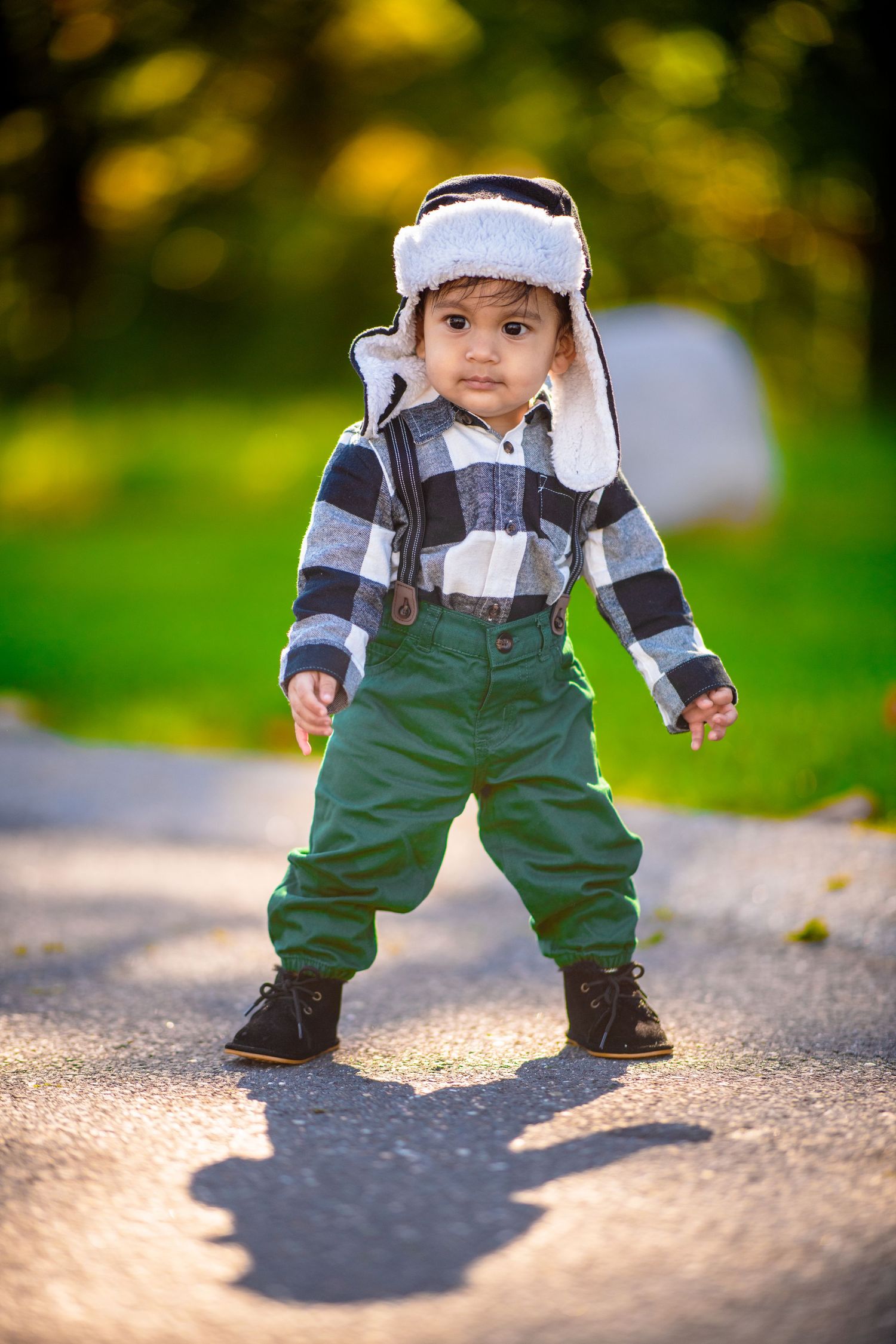 Small toddler in green overalls and white hat walking outdoors in autumn.