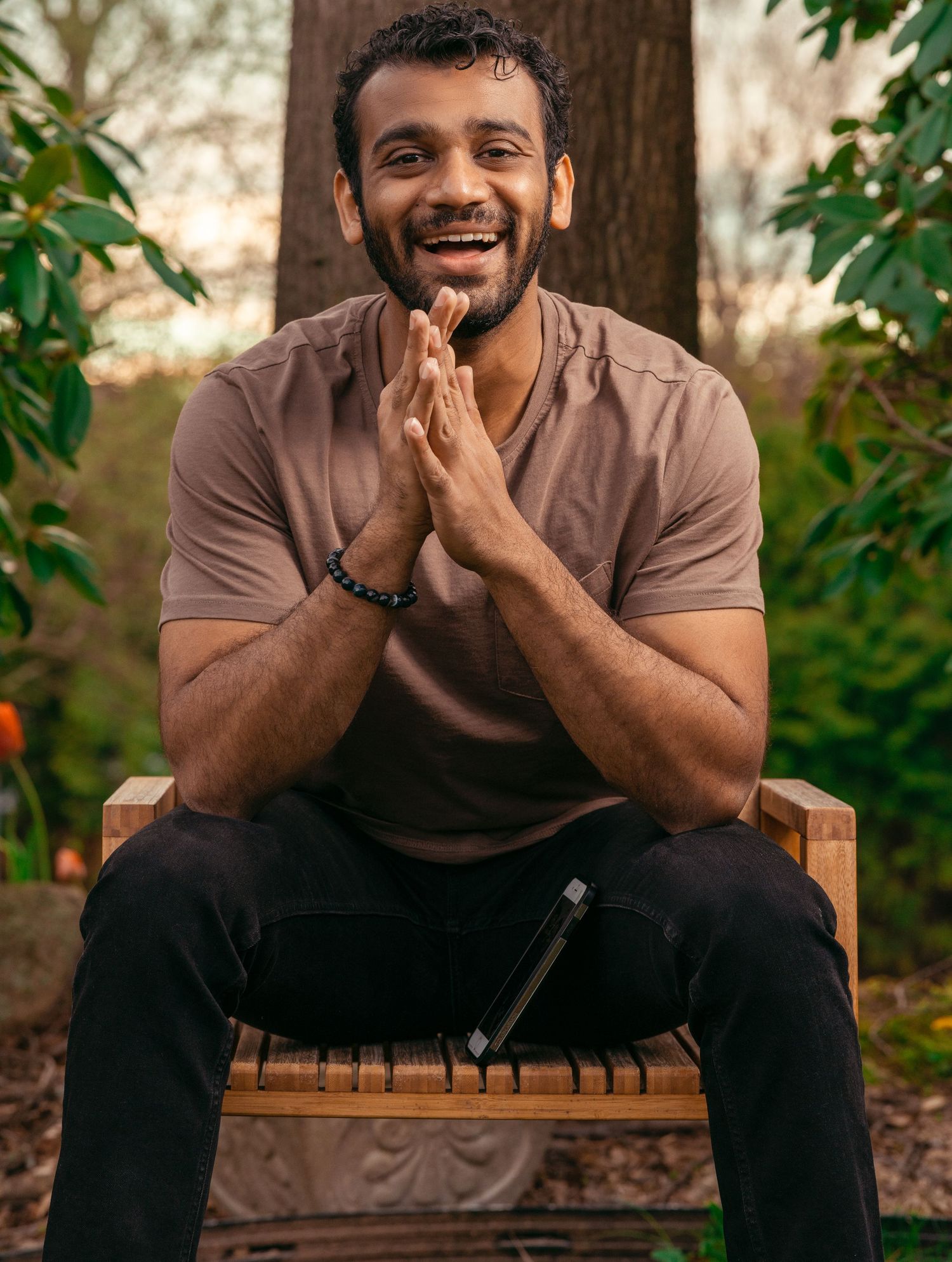 A person in a brown shirt sits outdoors on a wooden bench with clasped hands and a bright smile.