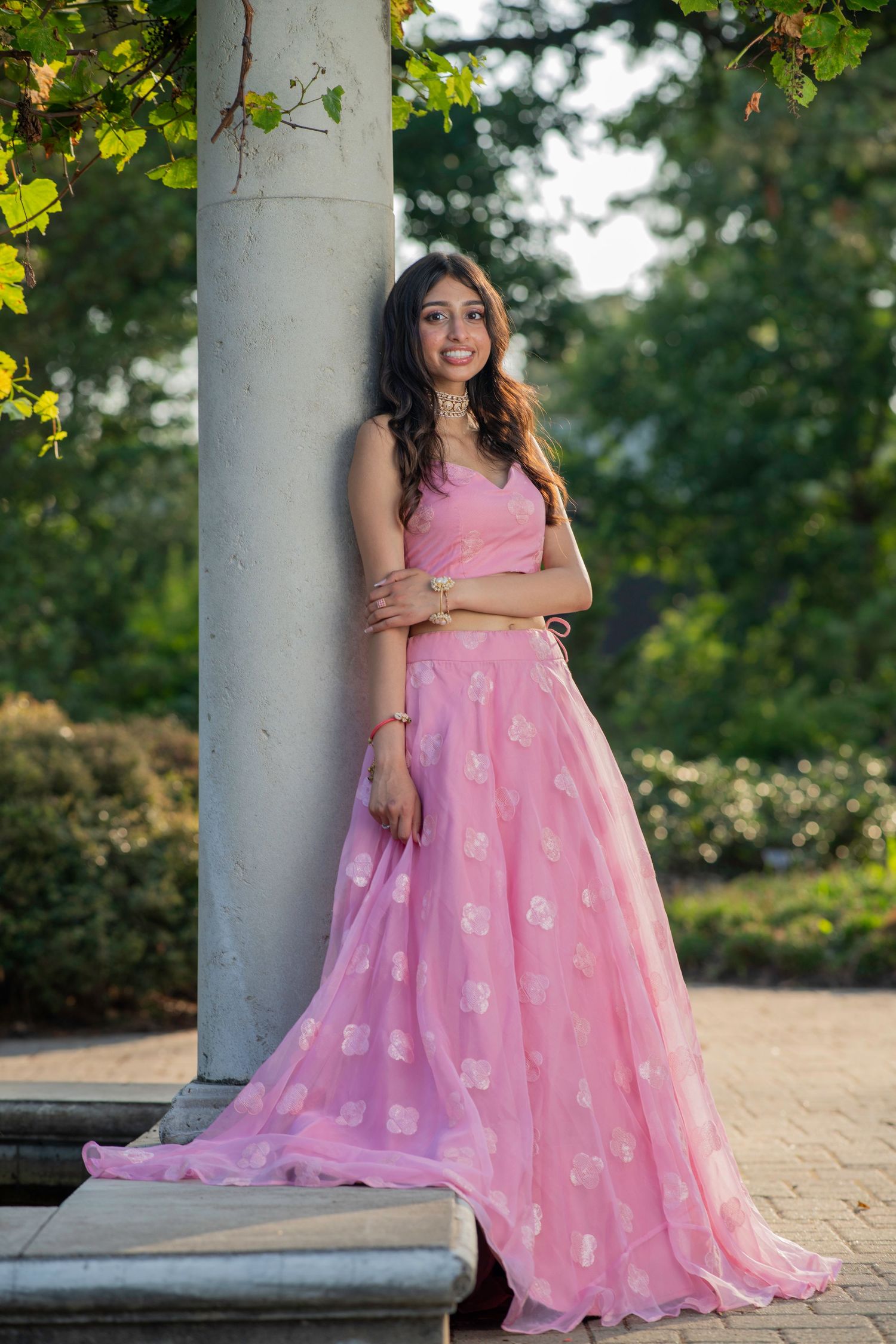 A flowing pink ball gown with delicate patterns is photographed against a garden backdrop with columns.