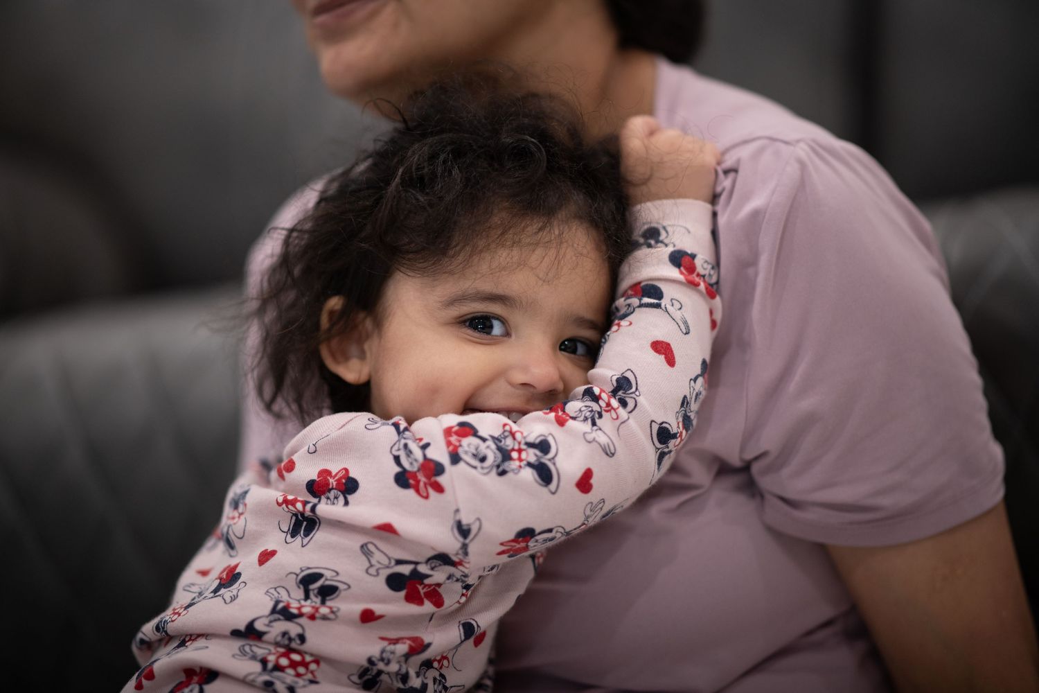 A small child in floral print pajamas hugs someone wearing a pink shirt while sitting on a gray couch.