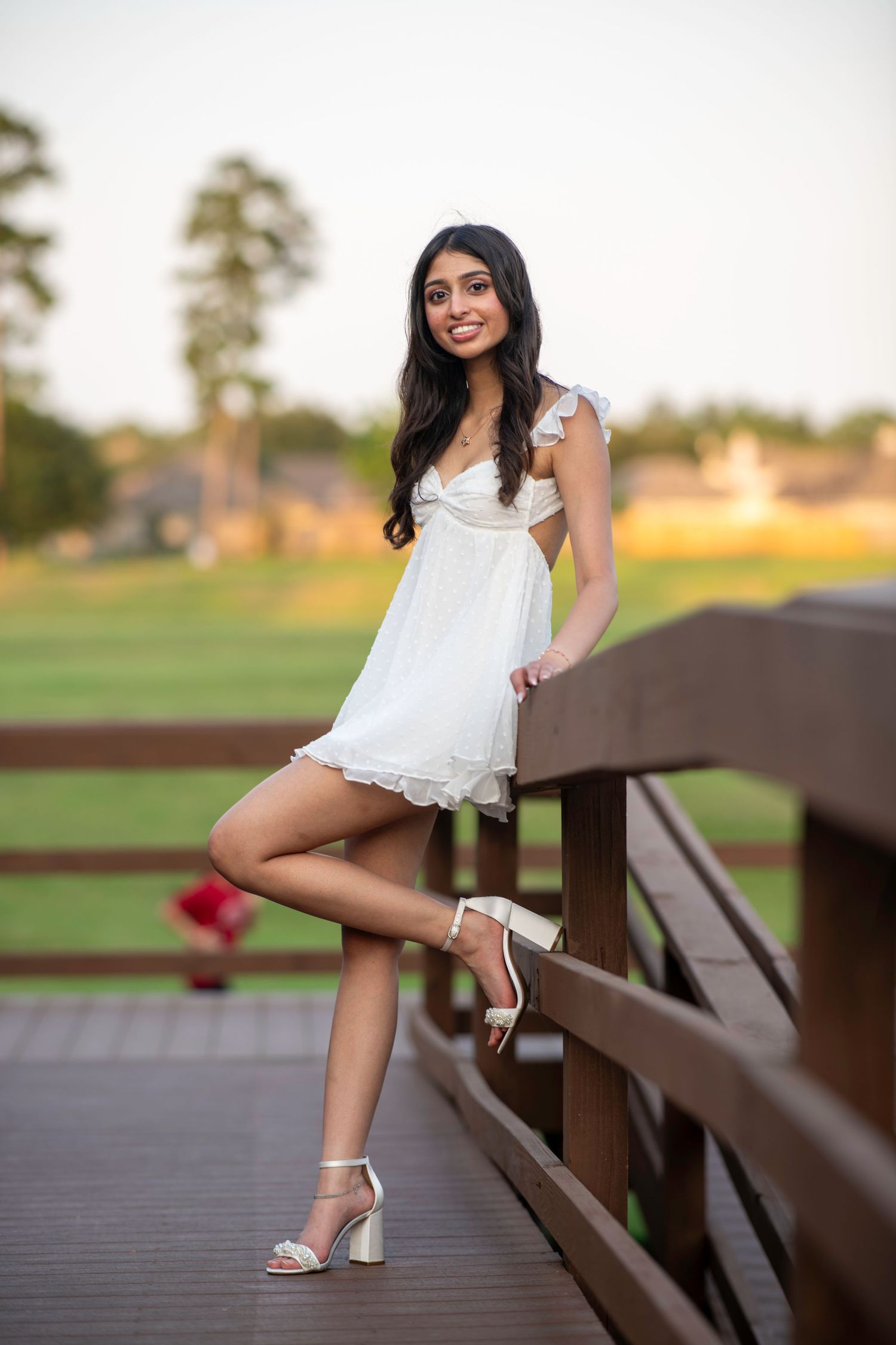 Woman in flowing white dress strikes elegant pose on wooden bridge during sunset photoshoot.