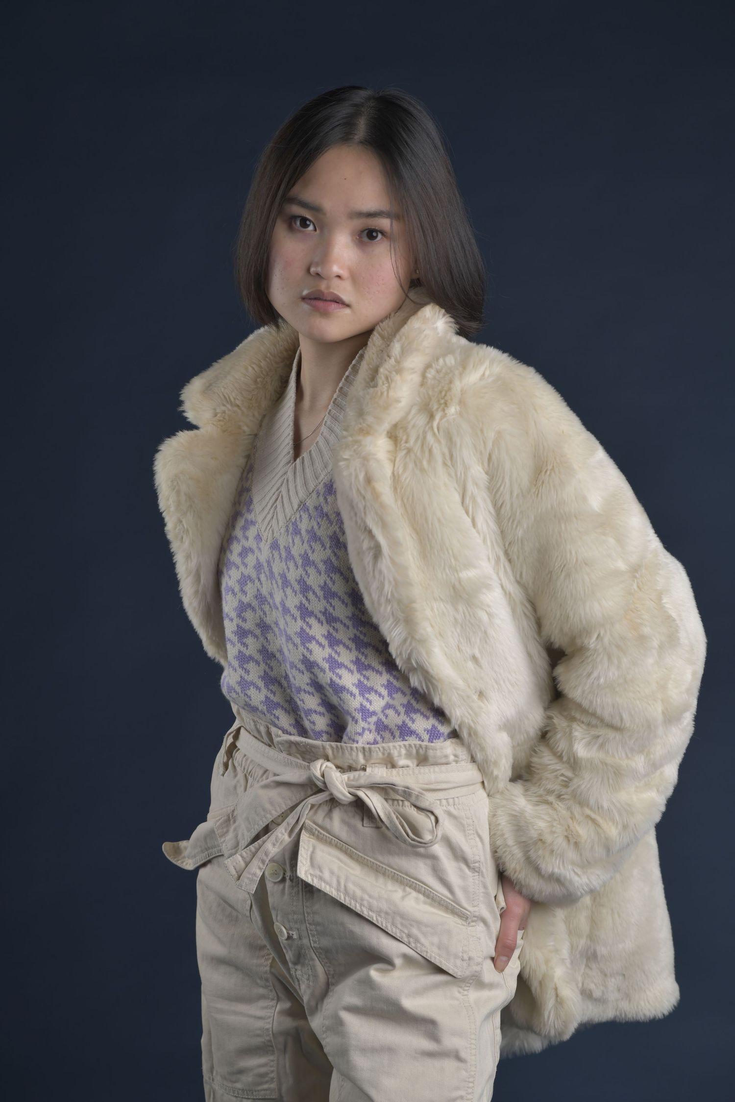 Fashion model wearing cream faux fur jacket with lavender patterned top and beige pants against dark background.