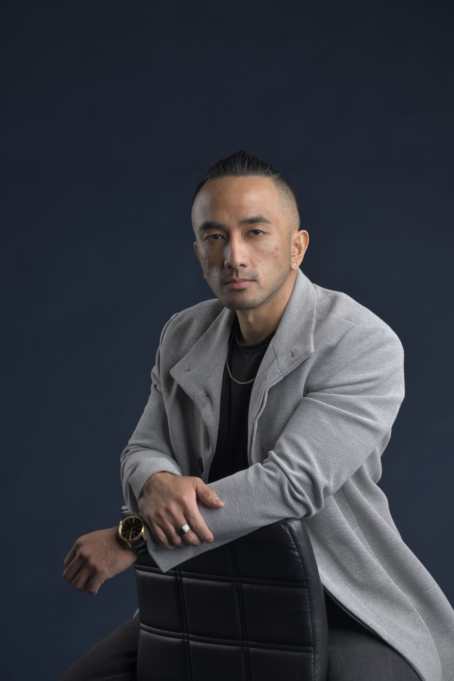 Professional studio portrait of a person in a light gray blazer and black shirt against a dark background.