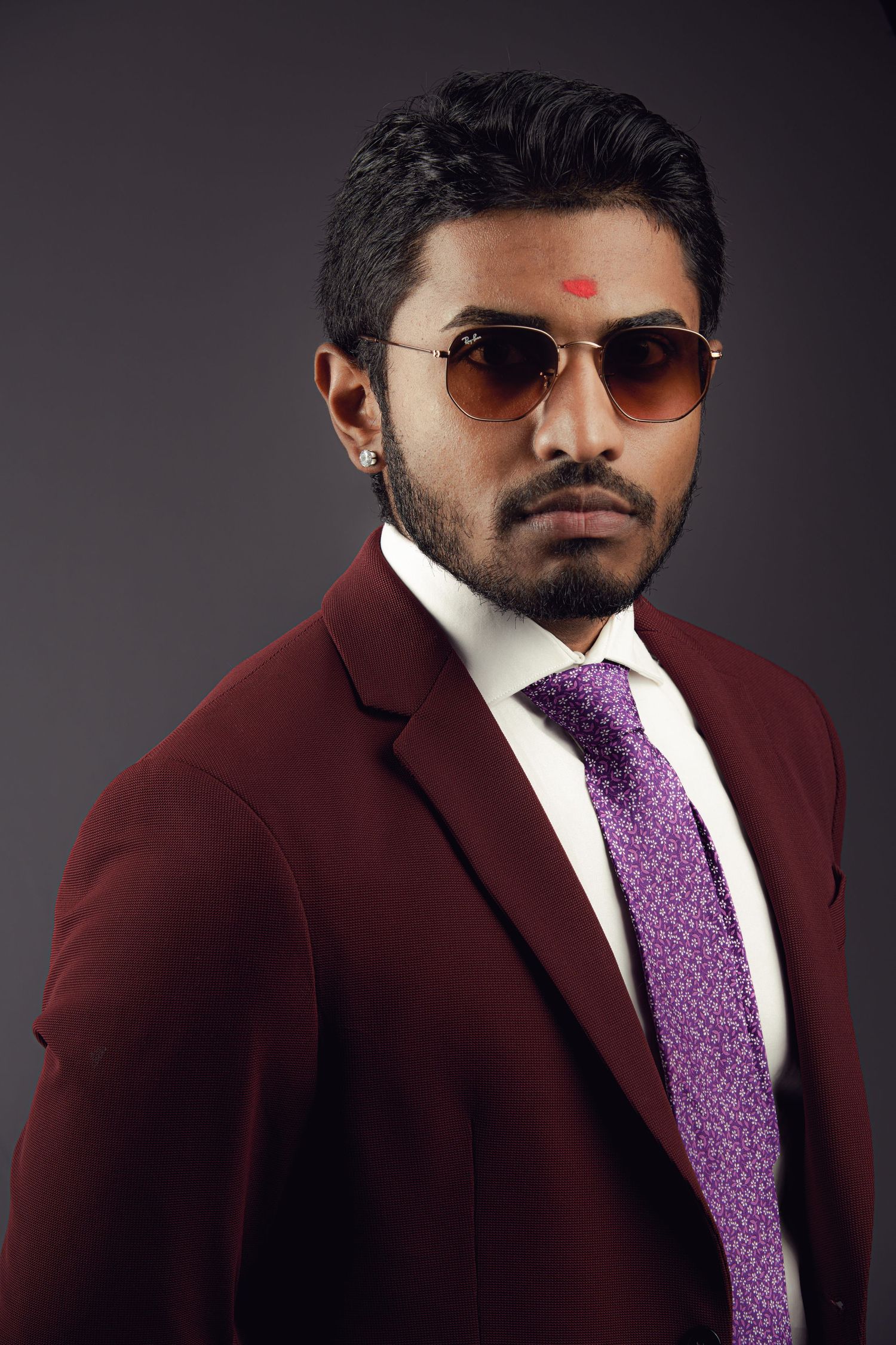 A person in a burgundy suit and purple tie poses against a dark background in a professional photo series.