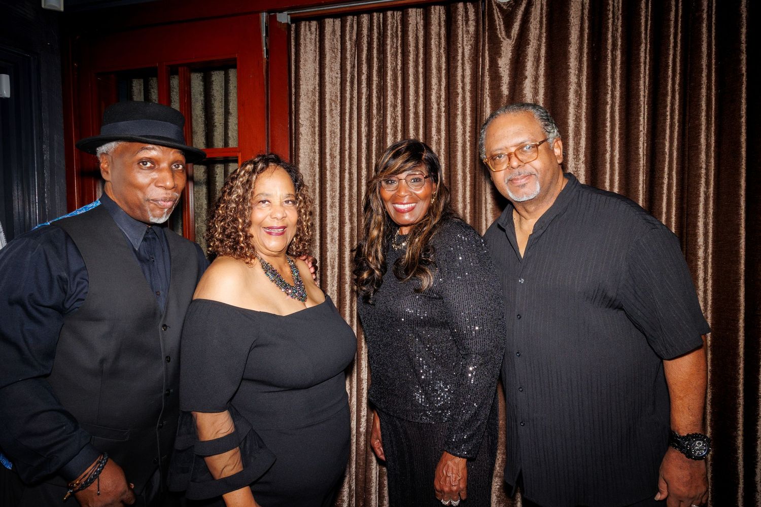 A group of people dressed in black gather for a social event against a dark curtained backdrop.