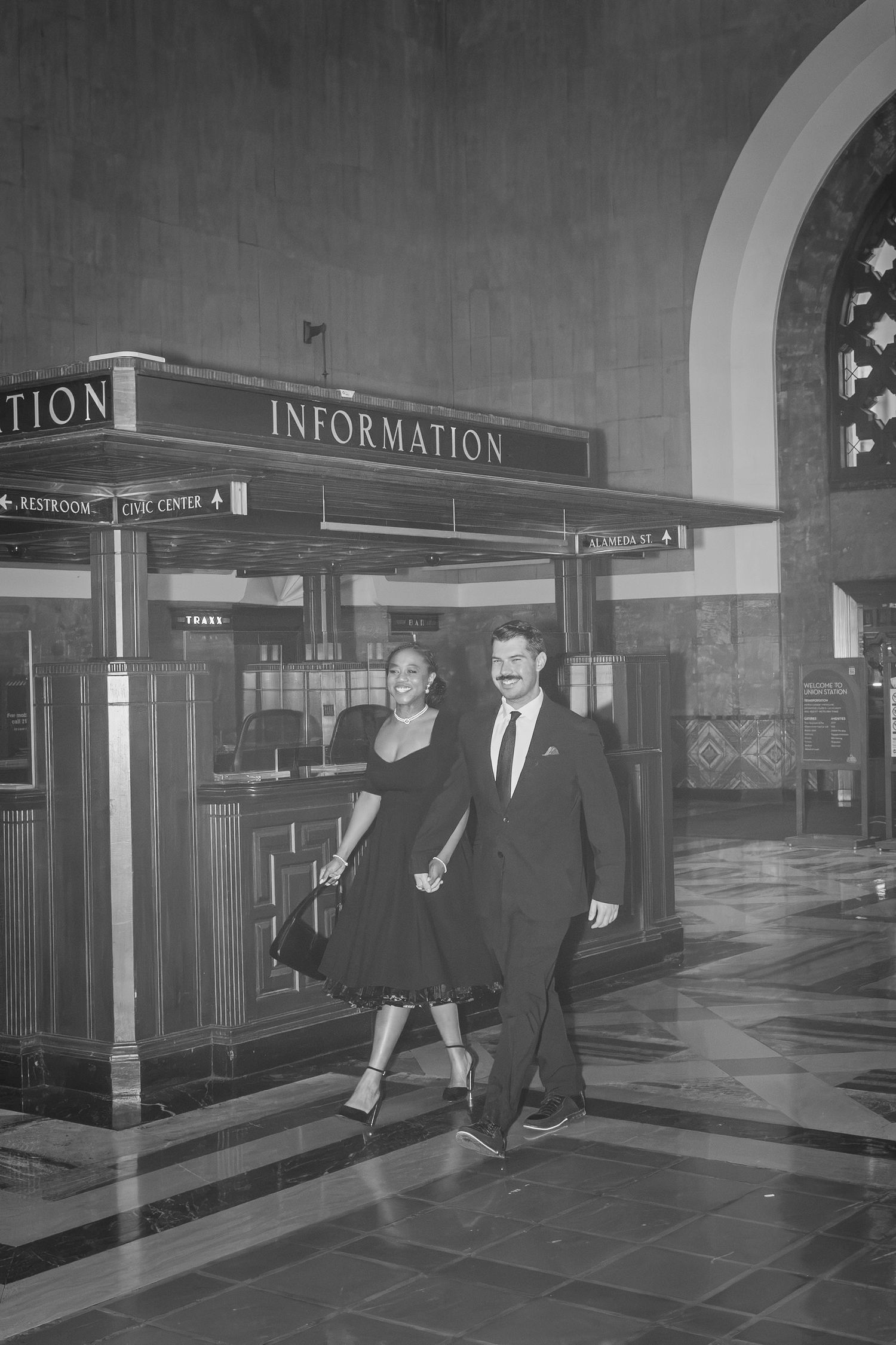 Vintage black and white photo of people walking past an information desk in a grand indoor space.