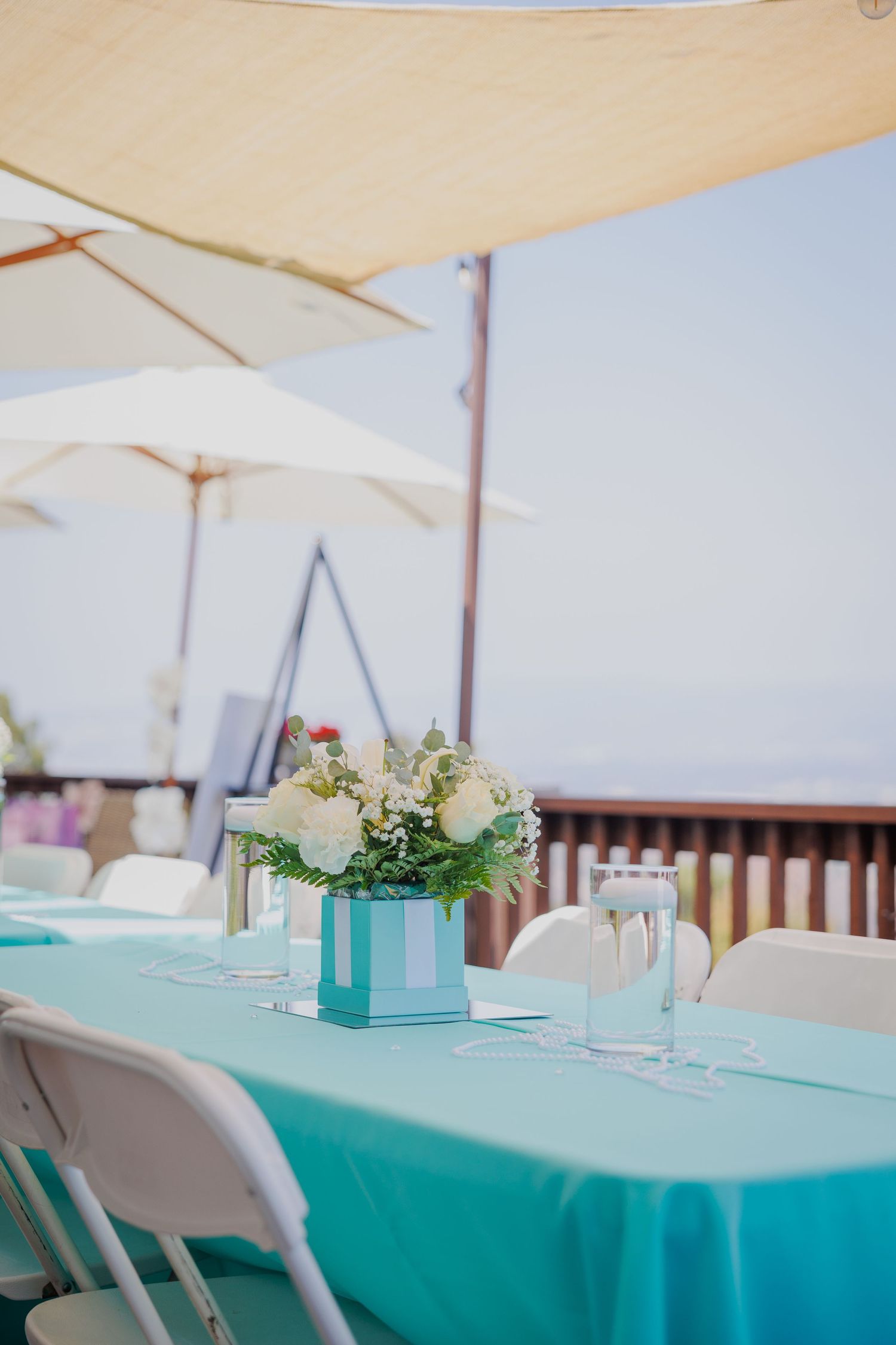 Elegant outdoor table setting with turquoise tablecloth and white floral centerpiece under shade umbrellas.