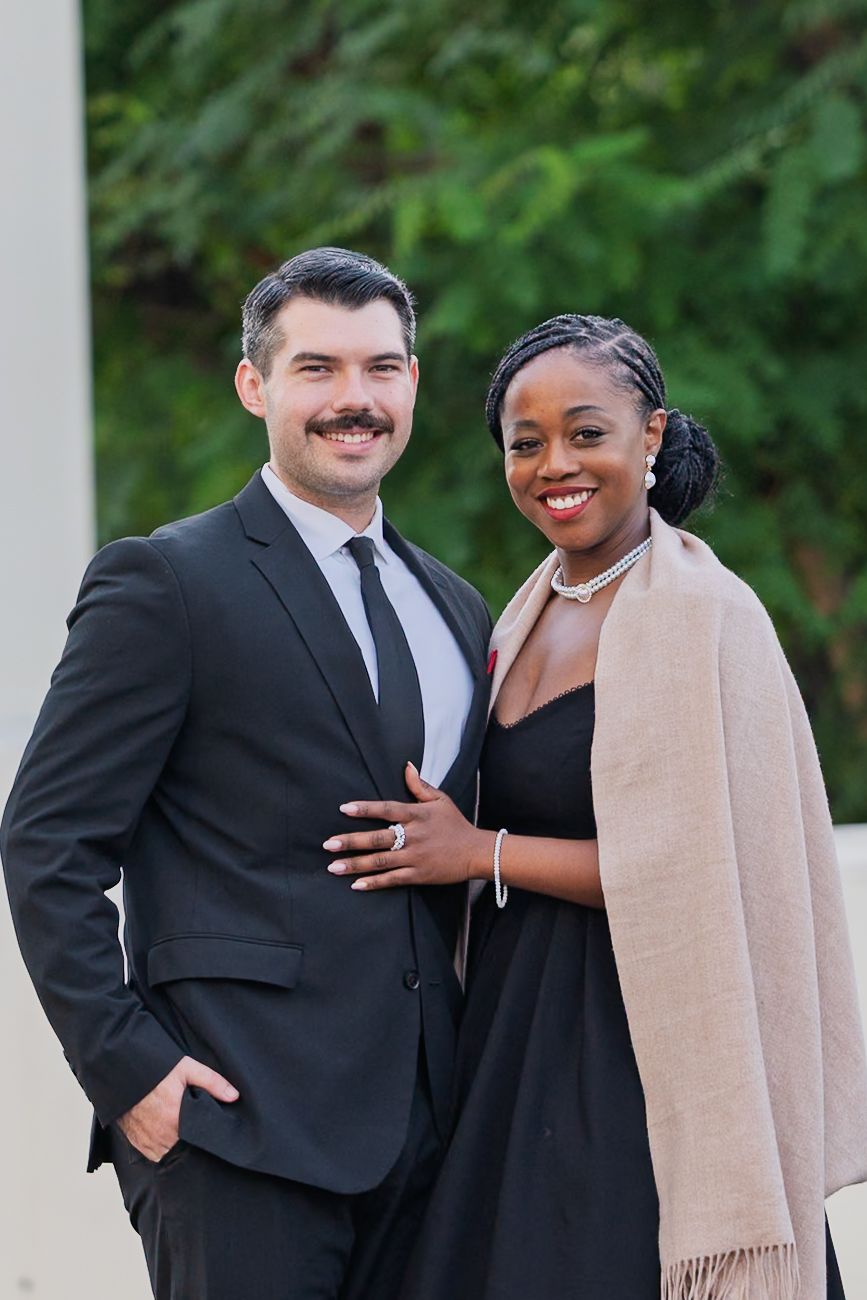 Couple in formal attire posing together outdoors with warm smiles.