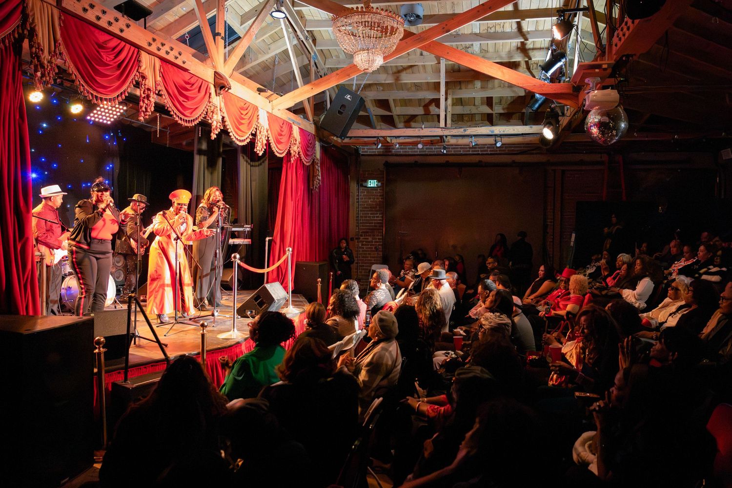 Wide shot of concert venue showing performers on stage with red curtains and full audience seating.