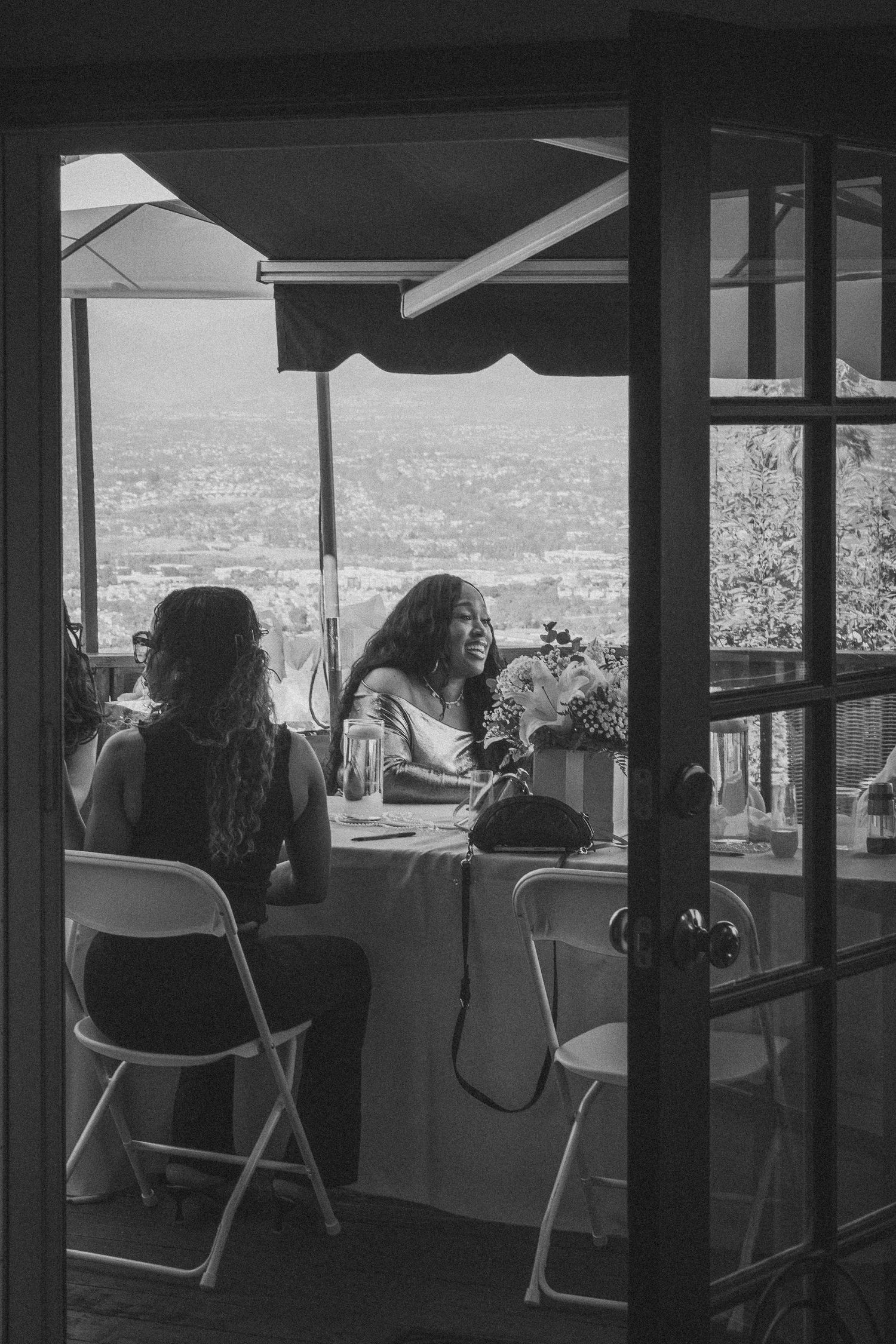 Two people enjoying drinks on a balcony overlooking a city view in black and white.