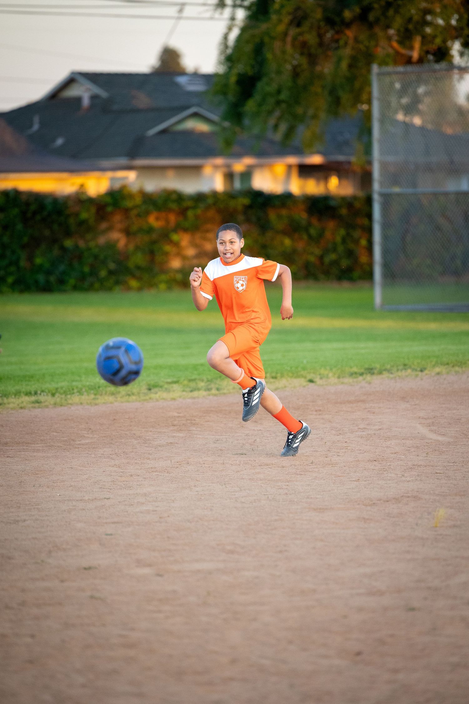 Soccer player in orange jersey kicking ball on dirt field at sunset.