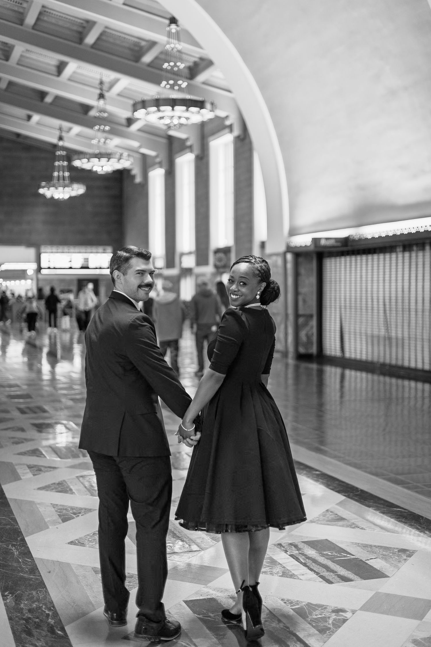 Black and white photograph showing couples walking through an elegant art deco style hallway with geometric floor patterns.