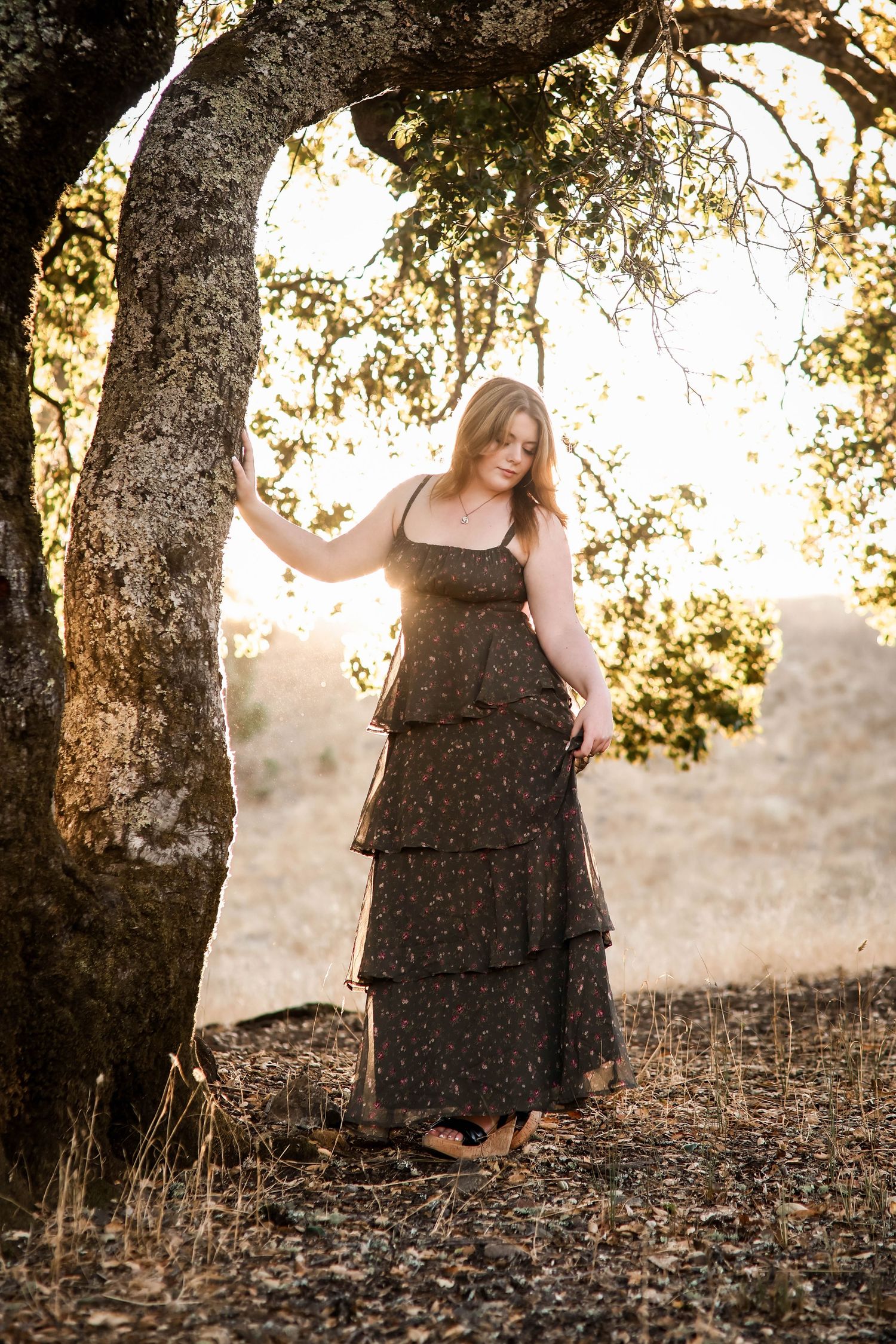 Woman in black tiered dress poses against tree during golden hour.