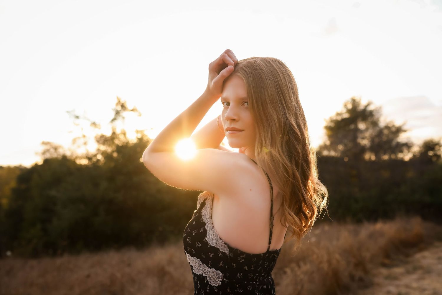 Sunkissed figure with long wavy hair stands in a lace top against golden sunset light in a natural outdoor setting.