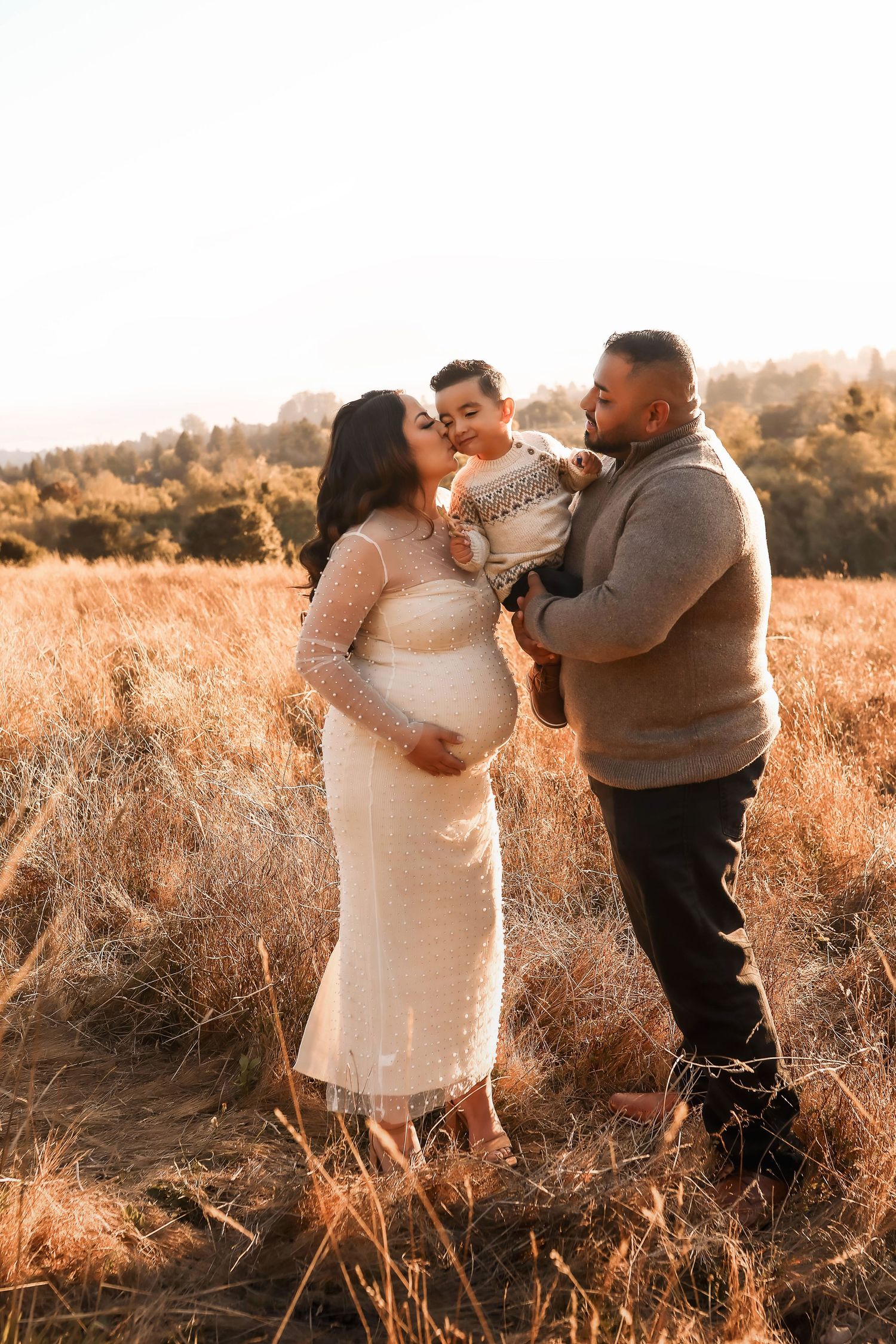 A family shares a sweet moment in golden sunlit field during outdoor sunset maternity photography session.