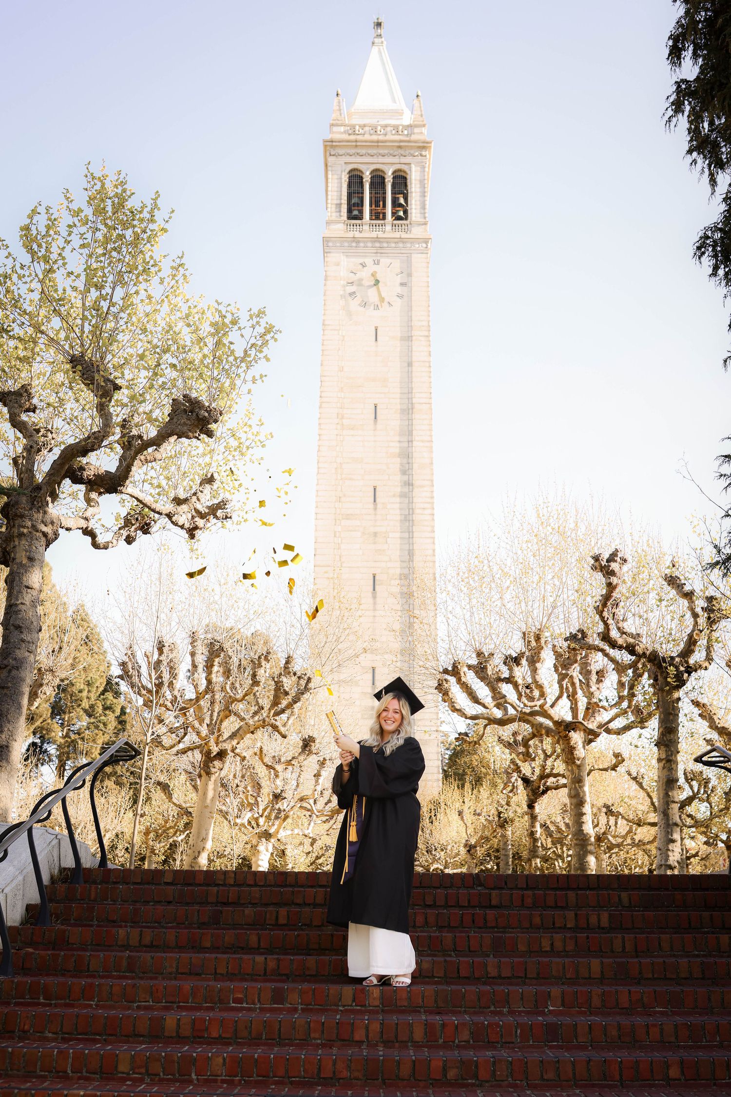 Confetti take flight in front of a tall church bell tower as silhouettes move on red brick steps below during a sunny day. Graduation.