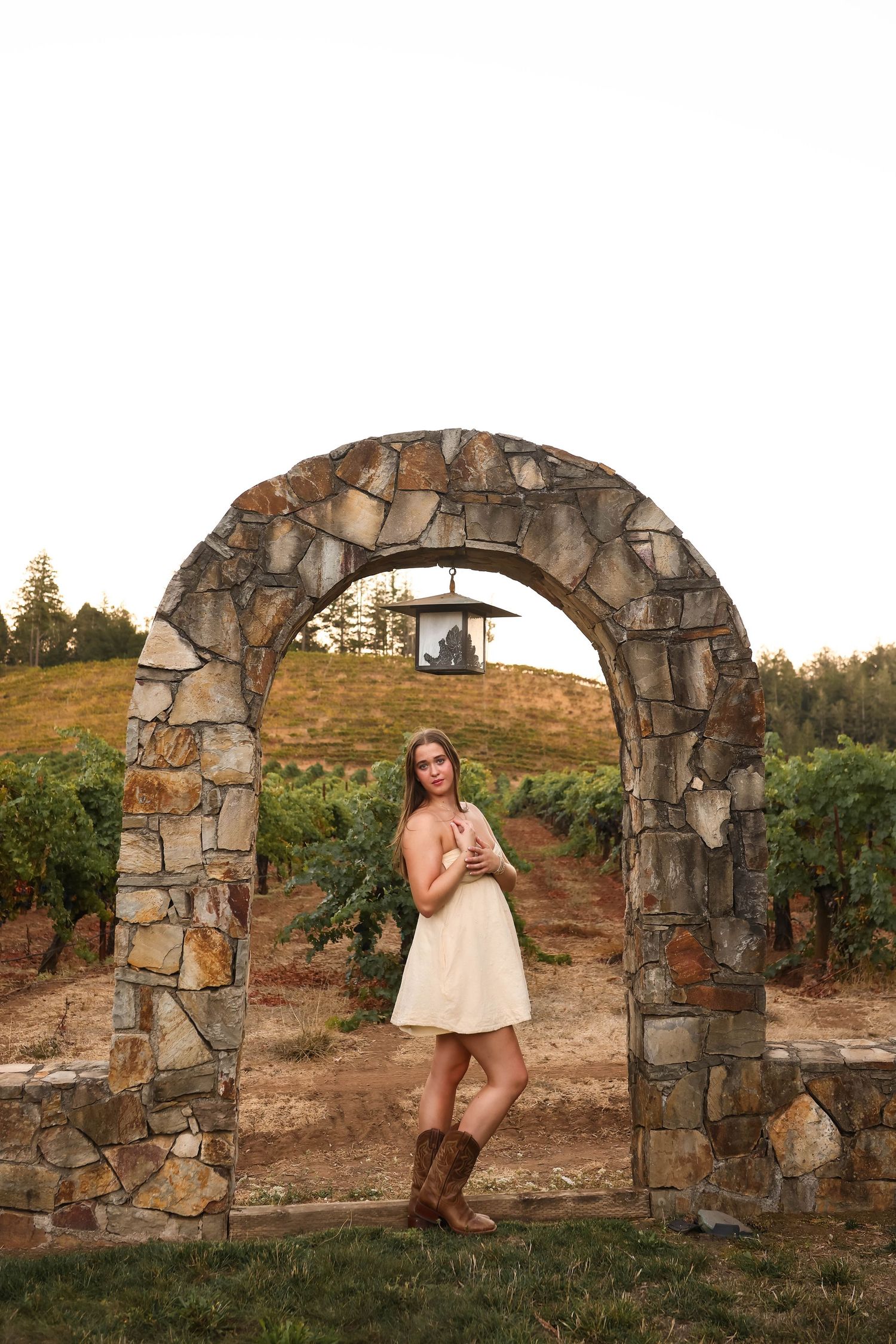 A stone archway stands in a vineyard at sunset with someone in a butter yellow dress and boots posing against it.