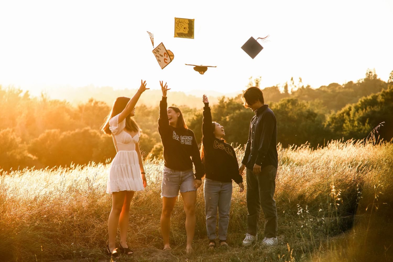 People walking along a sunset-lit field with tall golden grass and warm glowing backlight creating silhouettes. Graduation.