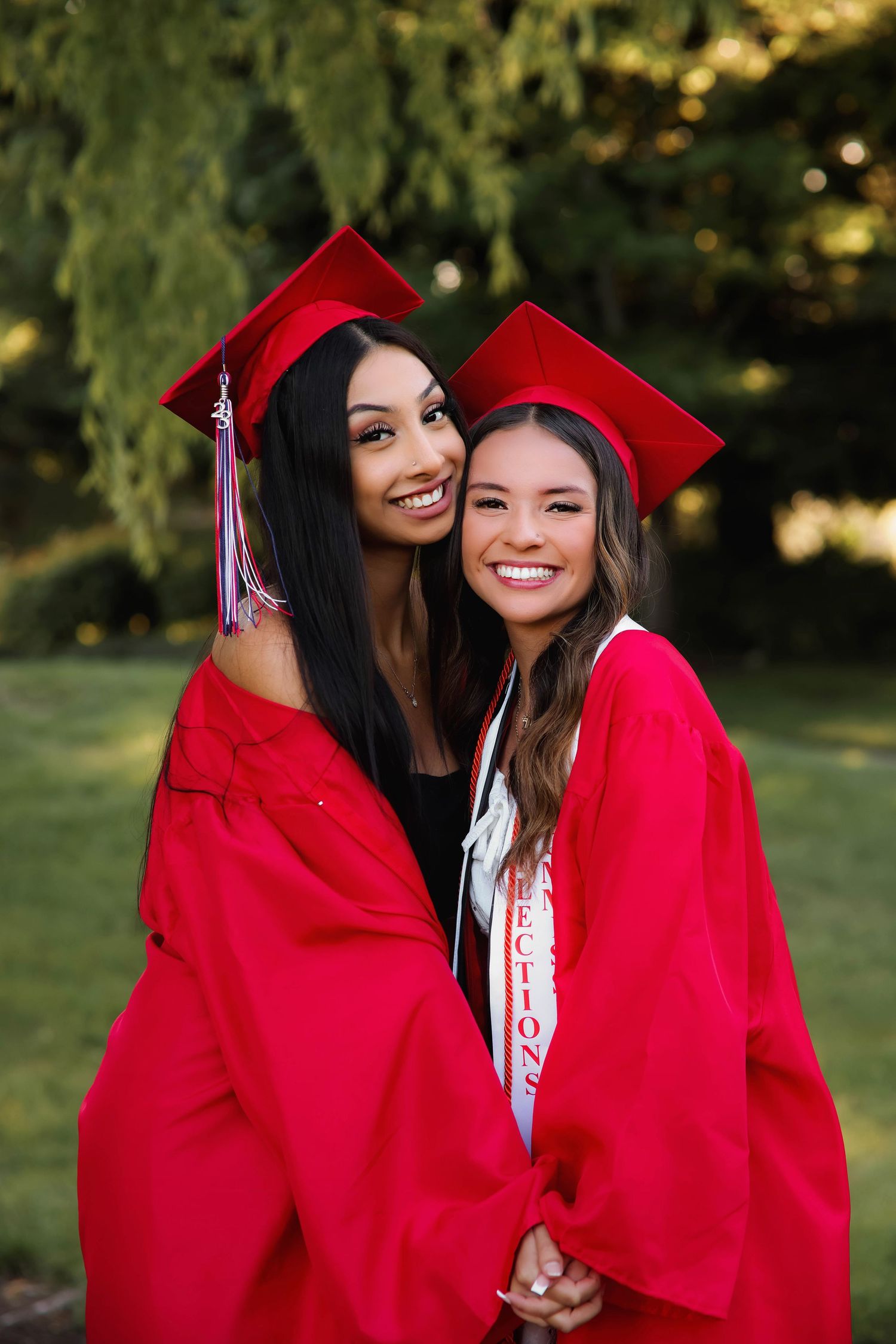 Two graduates wearing bright red caps and gowns smile together during outdoor commencement ceremony celebrations.