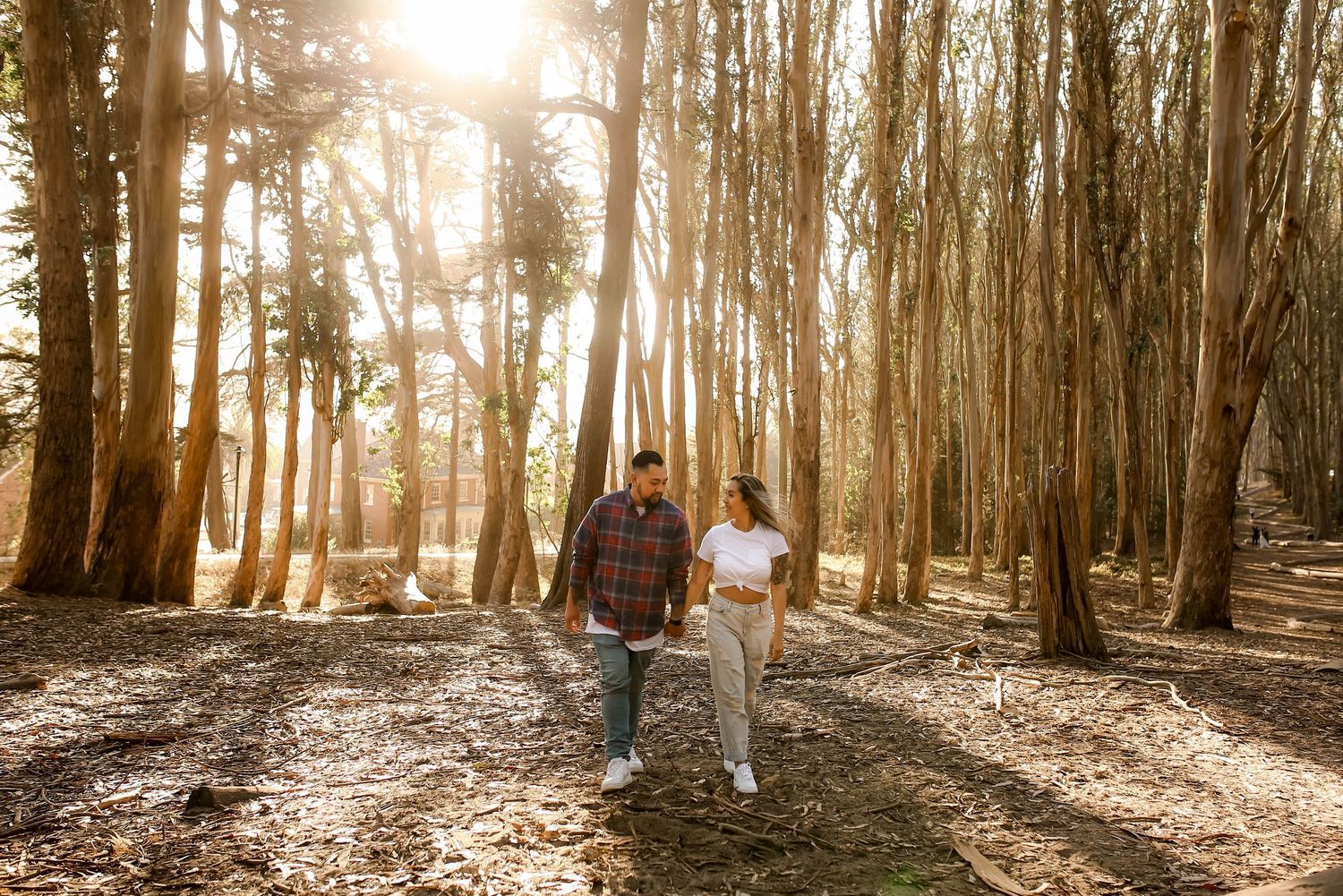 Sun rays filter through tall forest trees as a couple walks together on a leaf-covered path during golden hour.