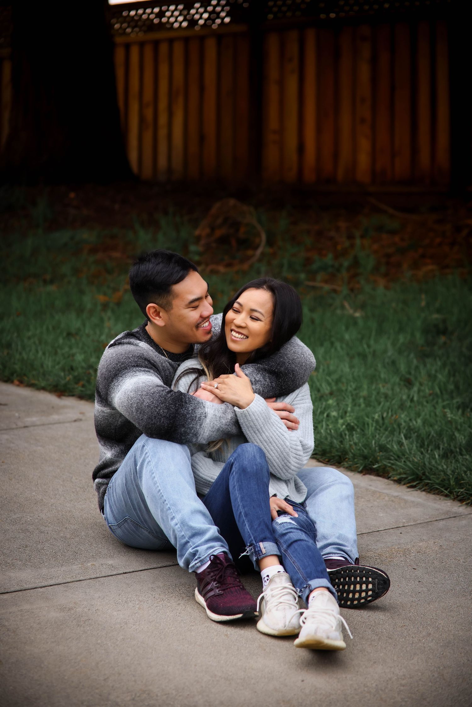 A couple sitting together on a concrete path share a joyful embrace while wearing casual sweaters and jeans.