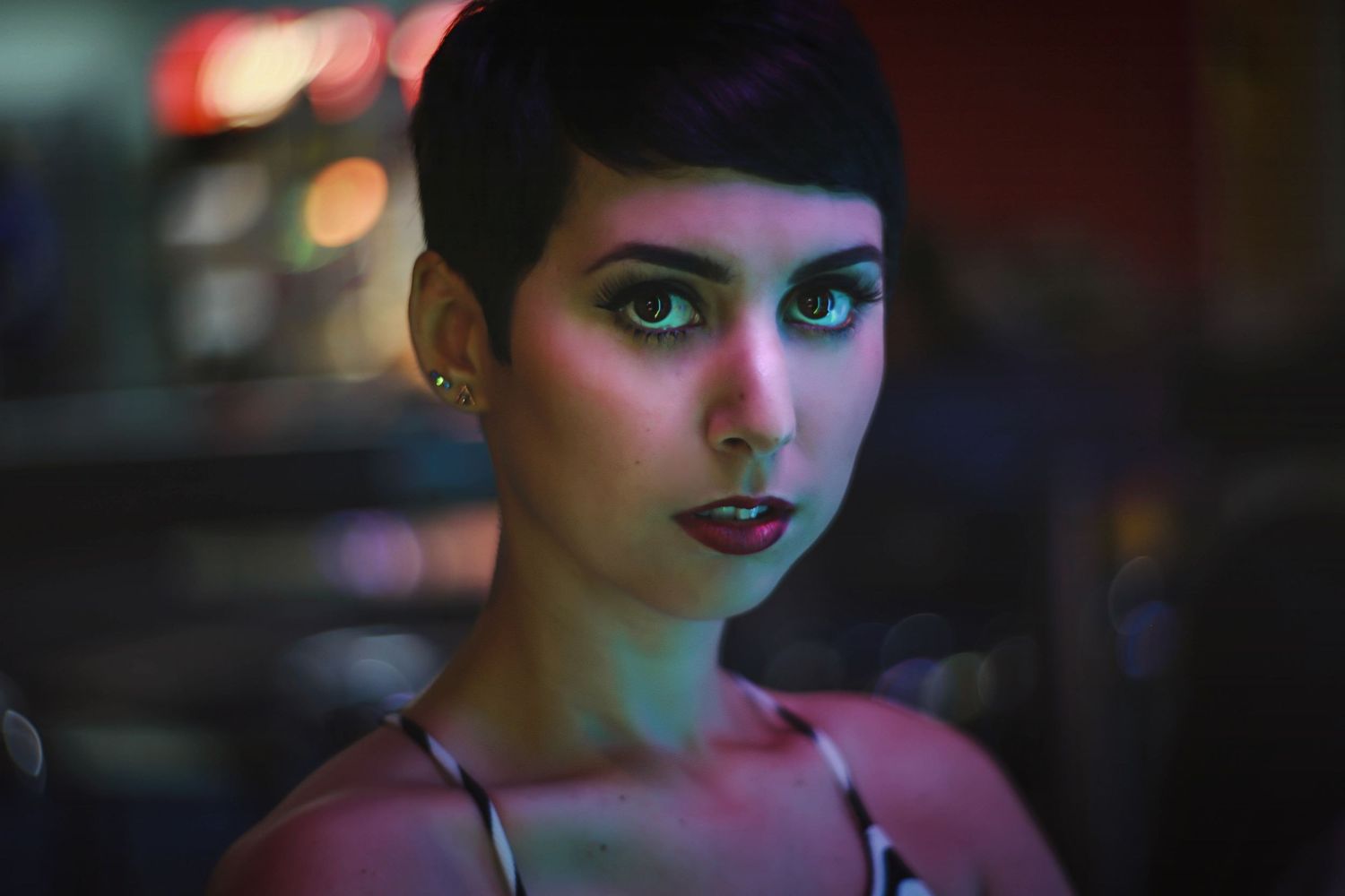 Dramatic portrait with short dark hair and vibrant makeup in moody pink and blue lighting.