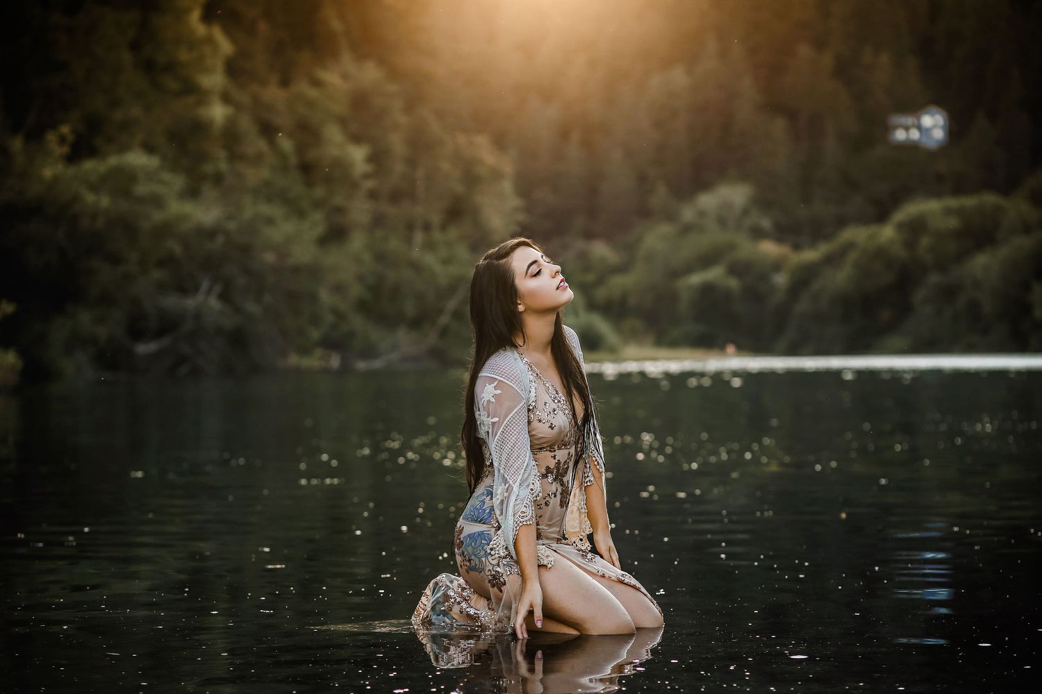 A person in light summer dress kneels at the edge of a serene river during golden hour with trees in the background.
