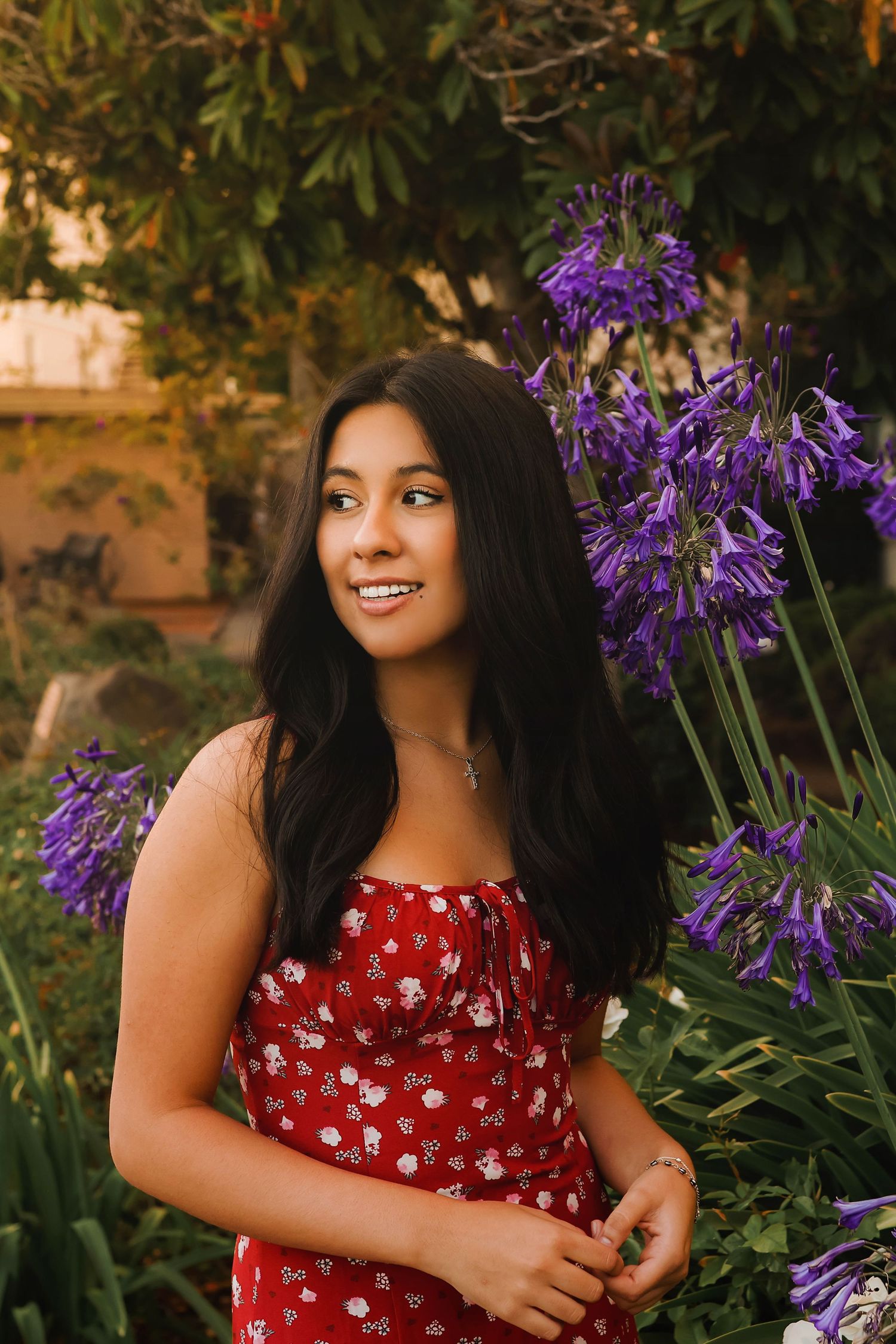 A young woman in a red polka dot dress smiles while standing next to vibrant purple agapanthus flowers in a garden.