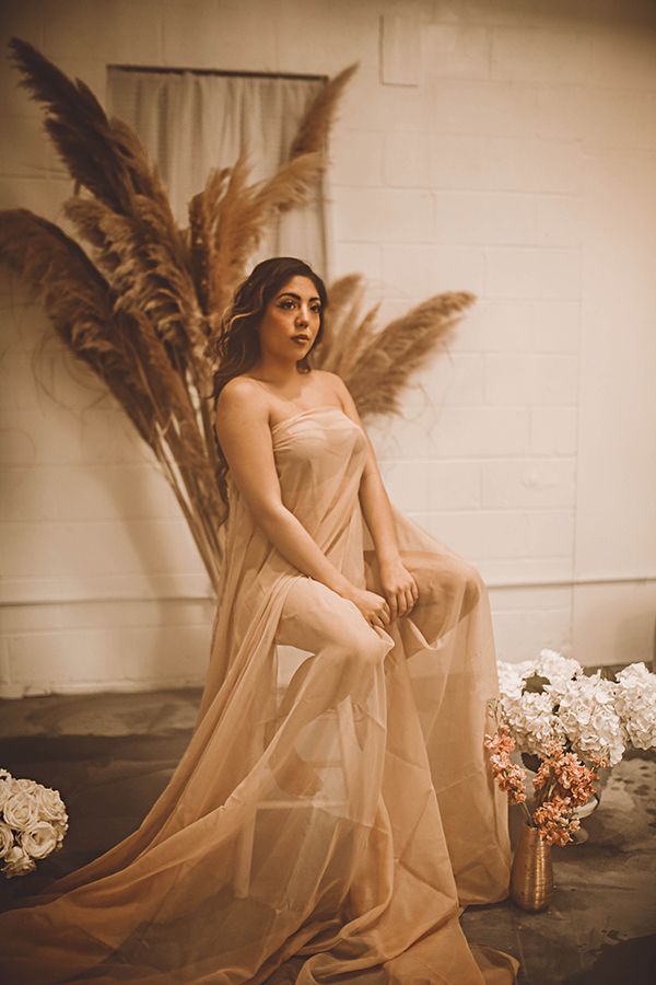 Ethereal photoshoot featuring feminine figure in flowing champagne-colored dress with dried pampas grass decor in sepia tones.