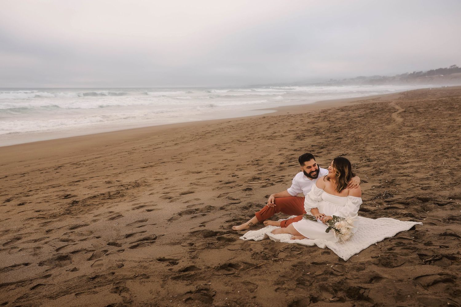Dreamy beachside photoshoot featuring a couple sitting on blanket at sunset with crashing waves in background.