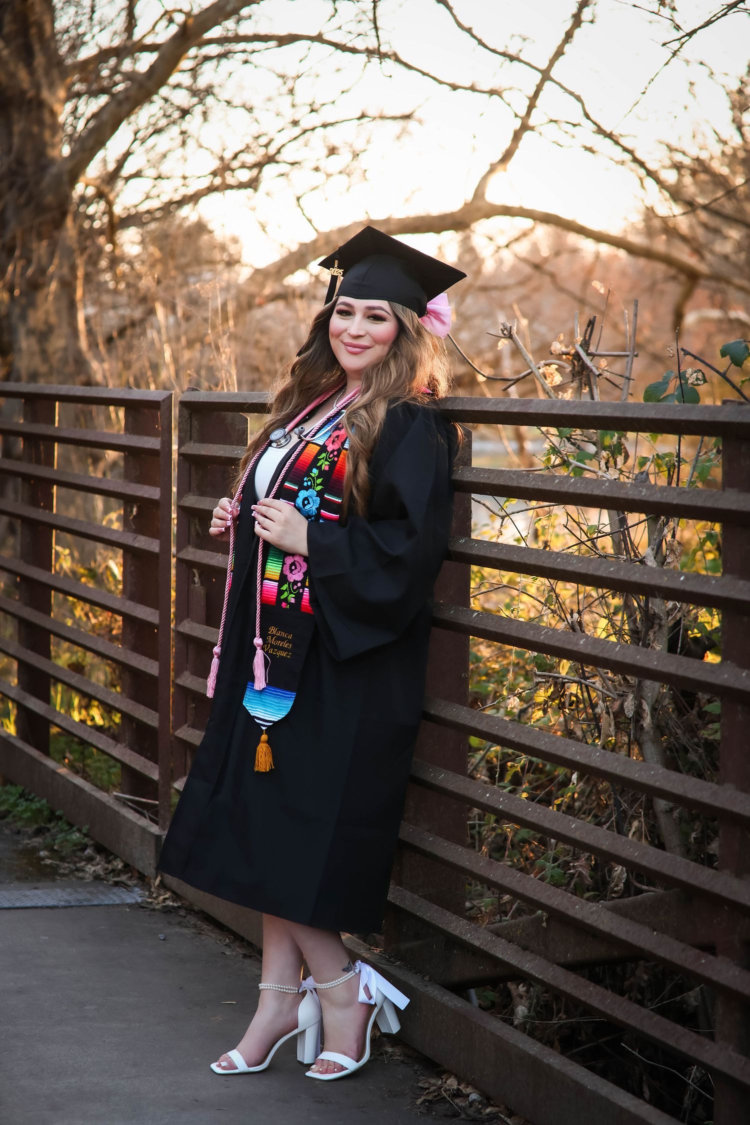 A graduate in traditional cap and gown poses on a wooden bridge at sunset during graduation photos.