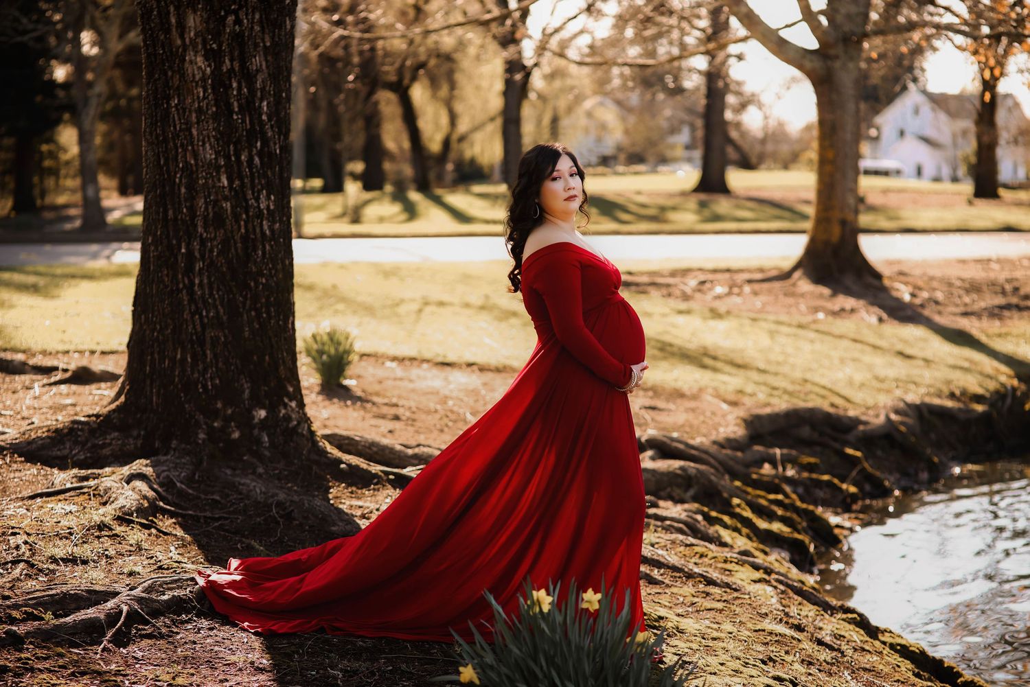 A woman in a red flowing dress moves dramatically in the wind near a tree and stream on a sunny autumn day. Maternity.