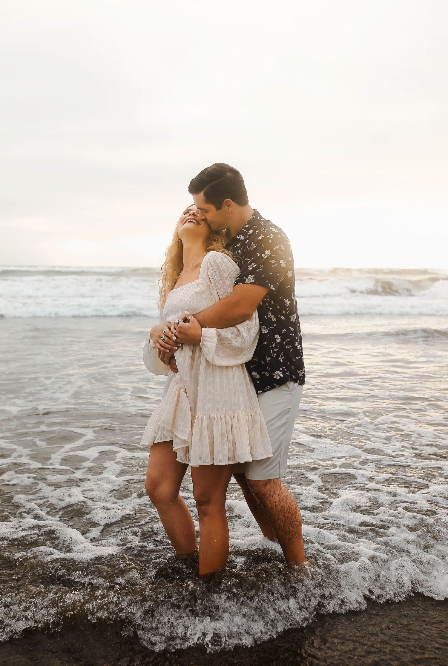 A romantic couple embraces while standing in the ocean waves at sunset on a sandy beach.