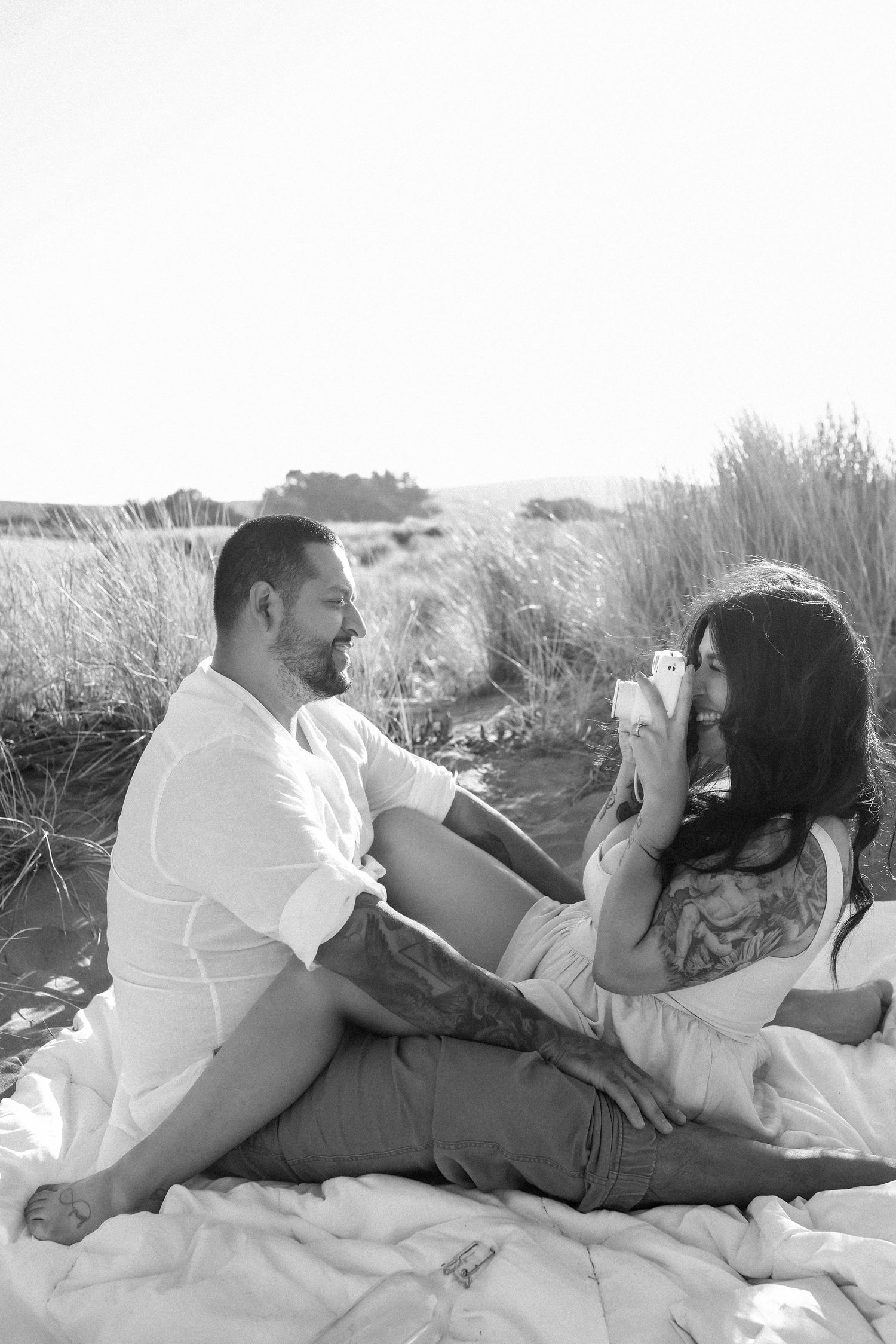 A romantic couple embraces and takes photos while lying in white sand dunes on a sunny day at the beach. Black and white photography.