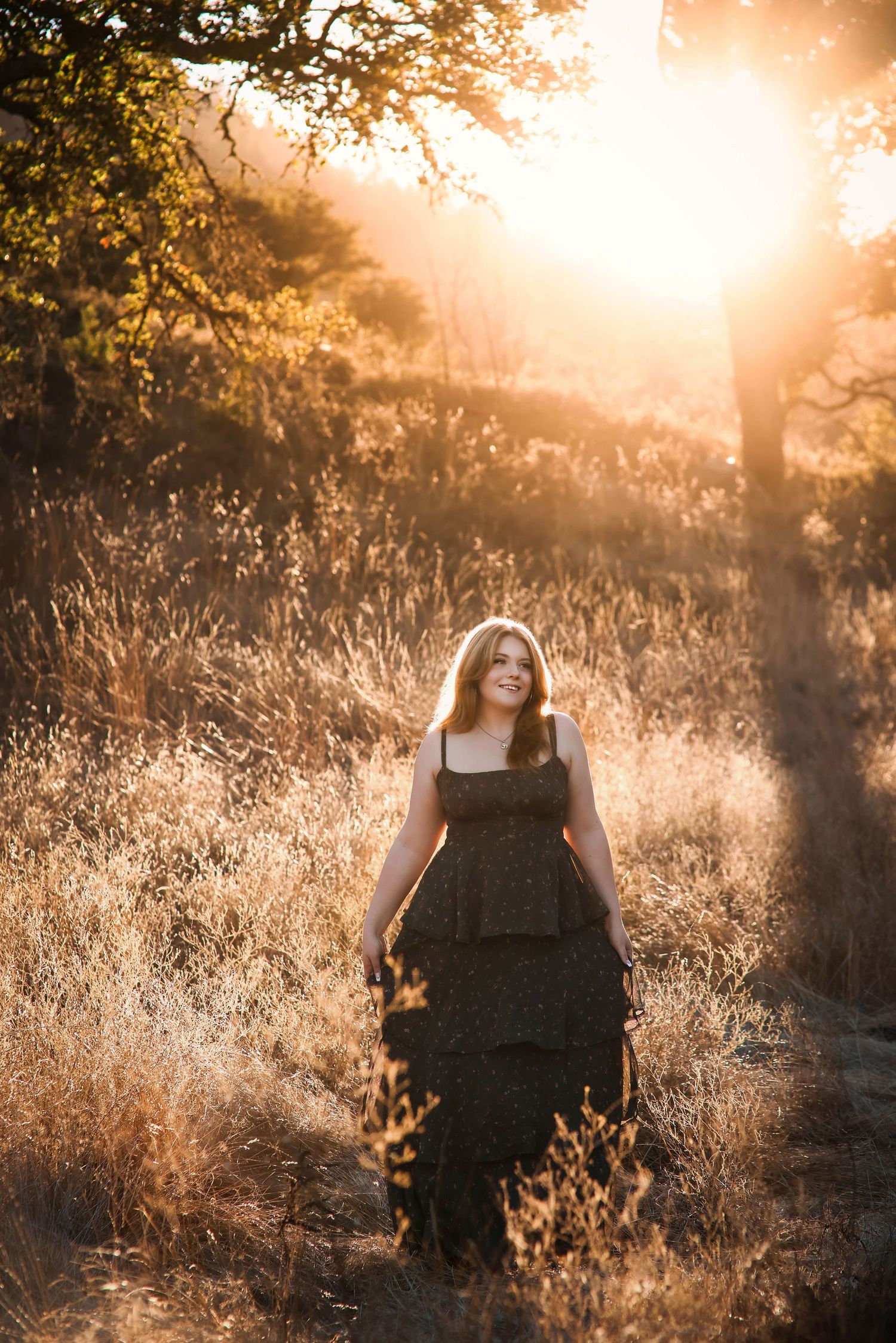A person poses in golden sunlit field with tall grass at sunset creating a dreamy ethereal atmosphere.