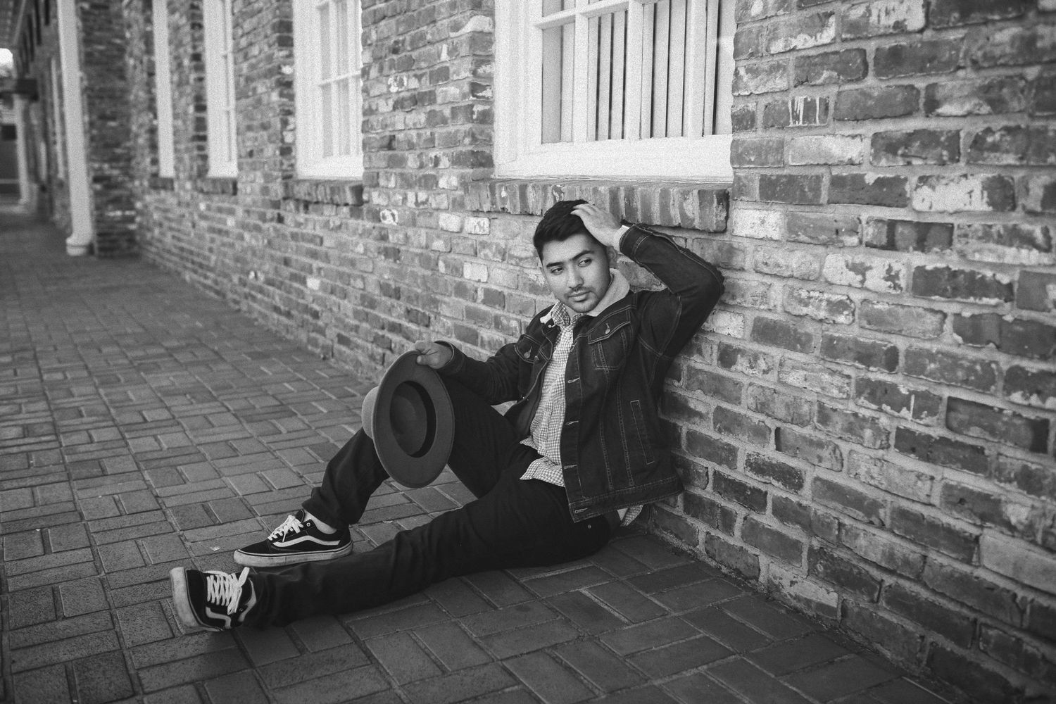 Black and white photo showing someone leaning against a horizontal brick wall while holding a hat and touching their head.