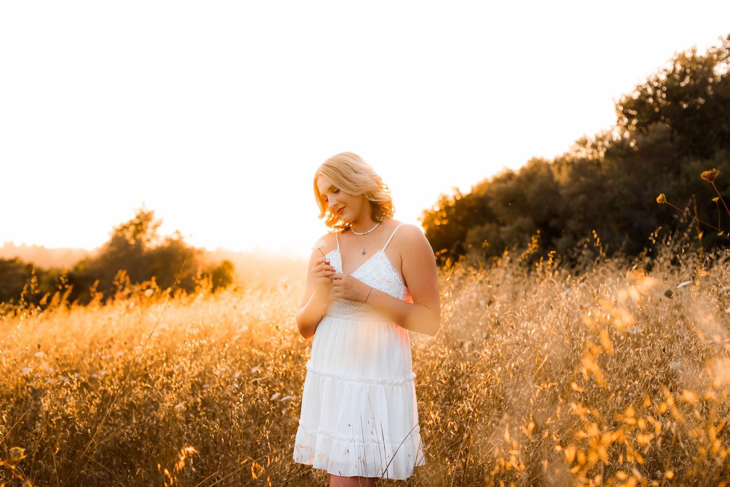 Woman in white dress standing in golden field at sunset with light glowing through tall grass.