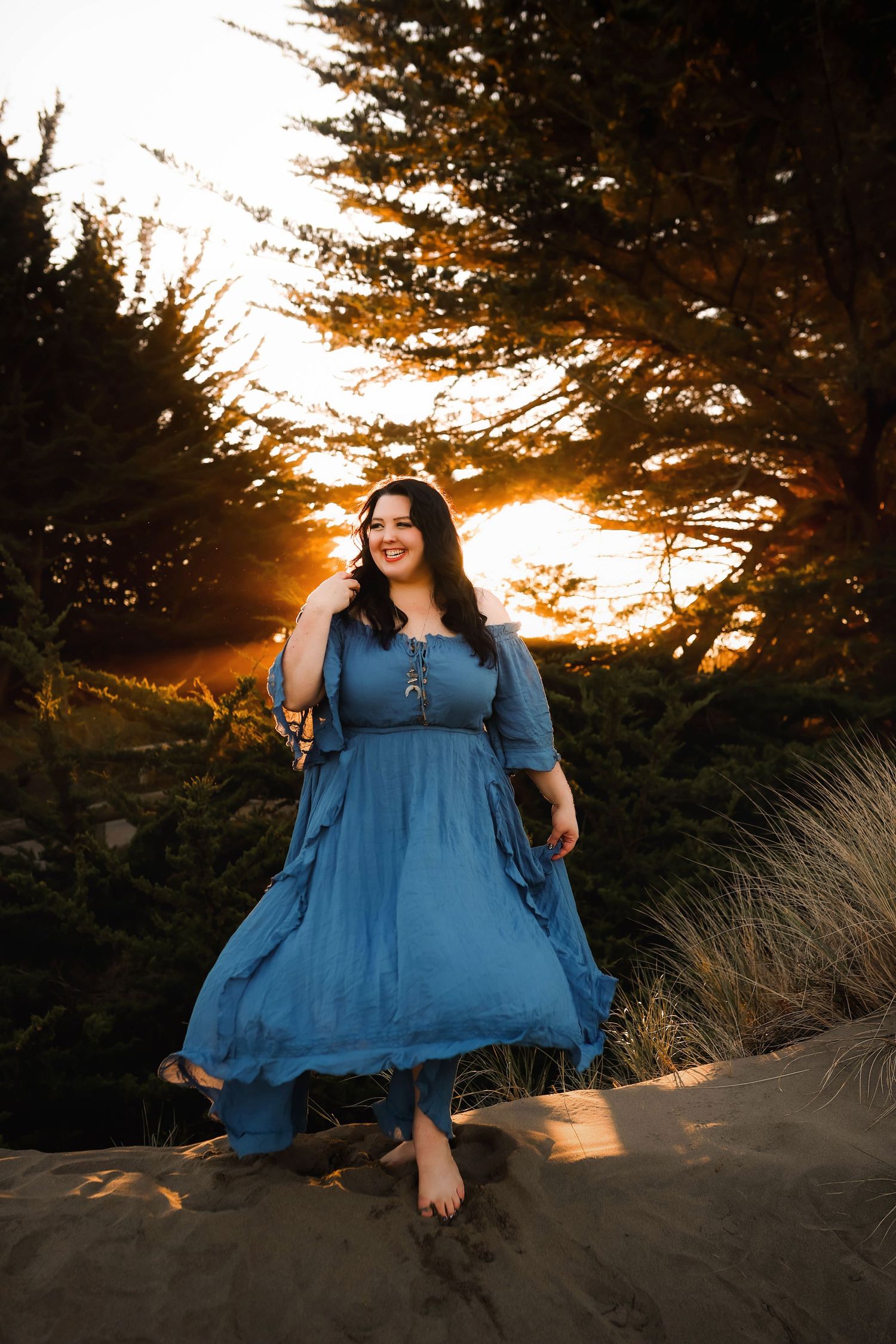 A person in a flowing blue dress stands barefoot on sand dunes at sunset while golden light filters through pine trees.