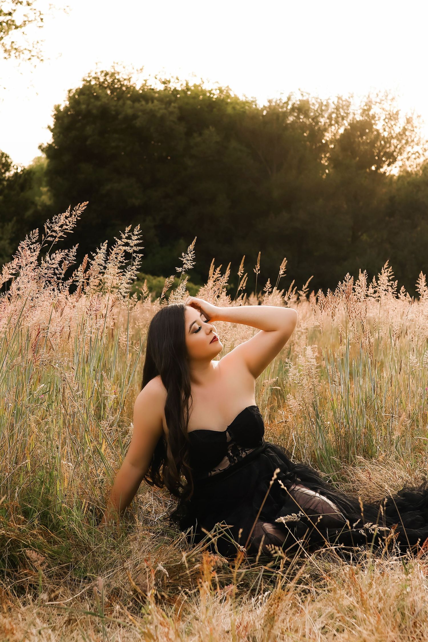 A figure in a black dress reclines gracefully in a sun-lit field of tall grass during golden hour.