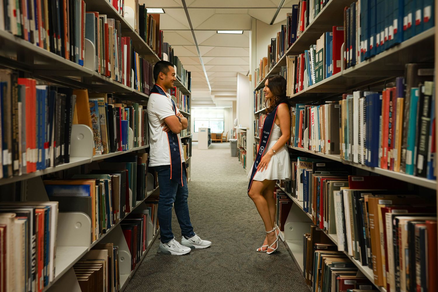 A couple standing in a library aisle with bookshelves stretching into the distance. Graduation.
