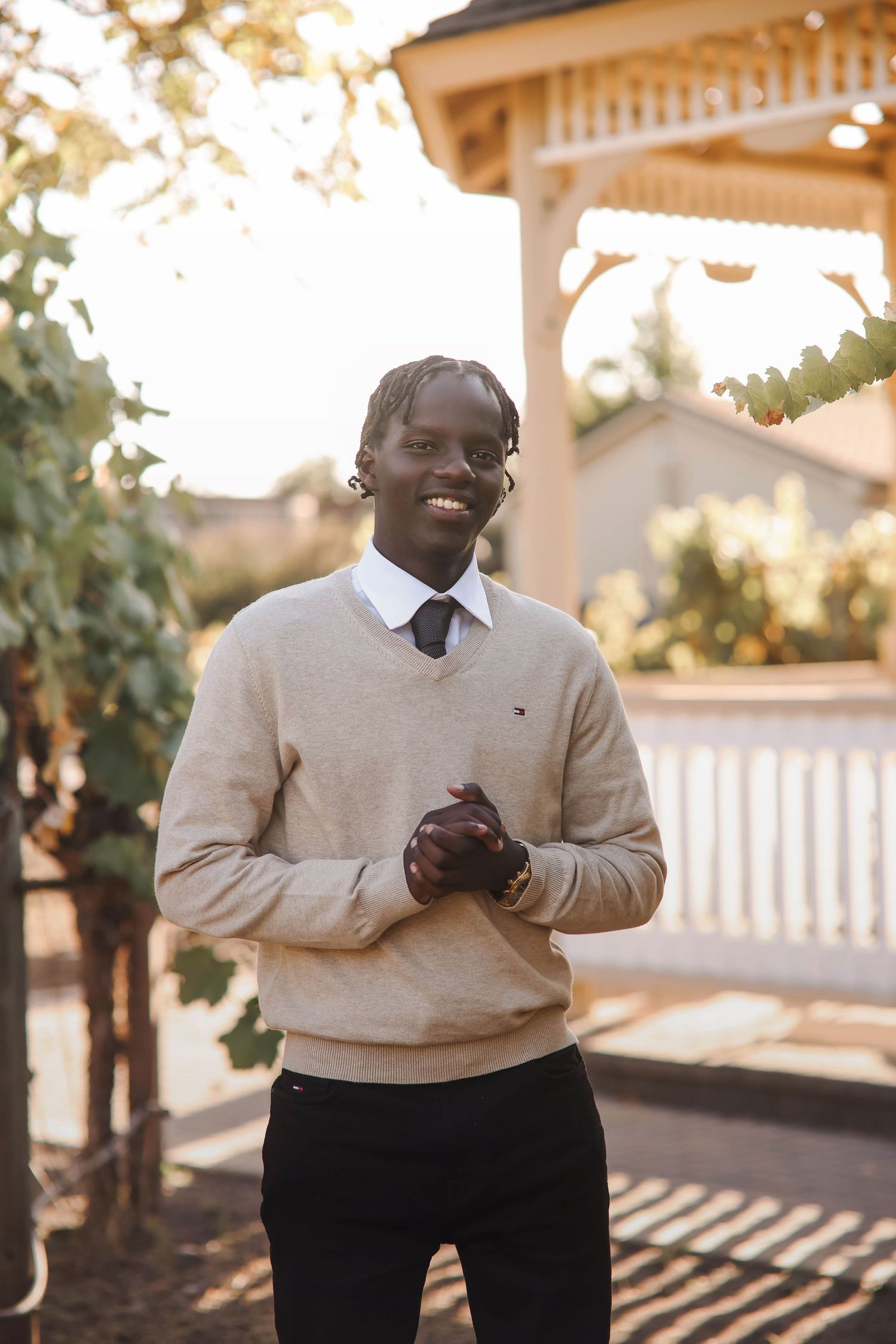 Someone wearing a beige sweater and white shirt with tie smiles while standing in front of a gazebo on a sunny day.