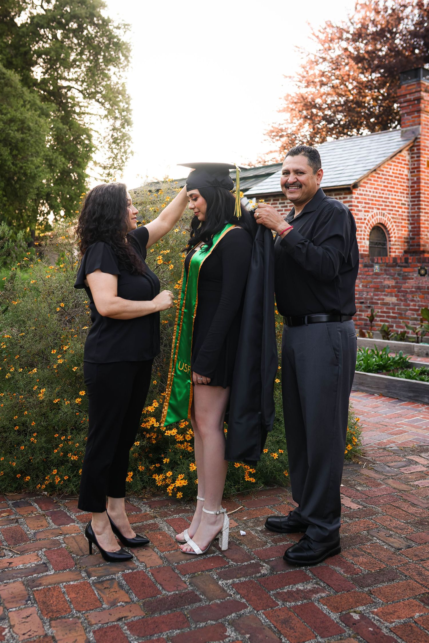 A graduation celebration takes place on a brick path outside a church with flowering bushes and fall foliage in the background.