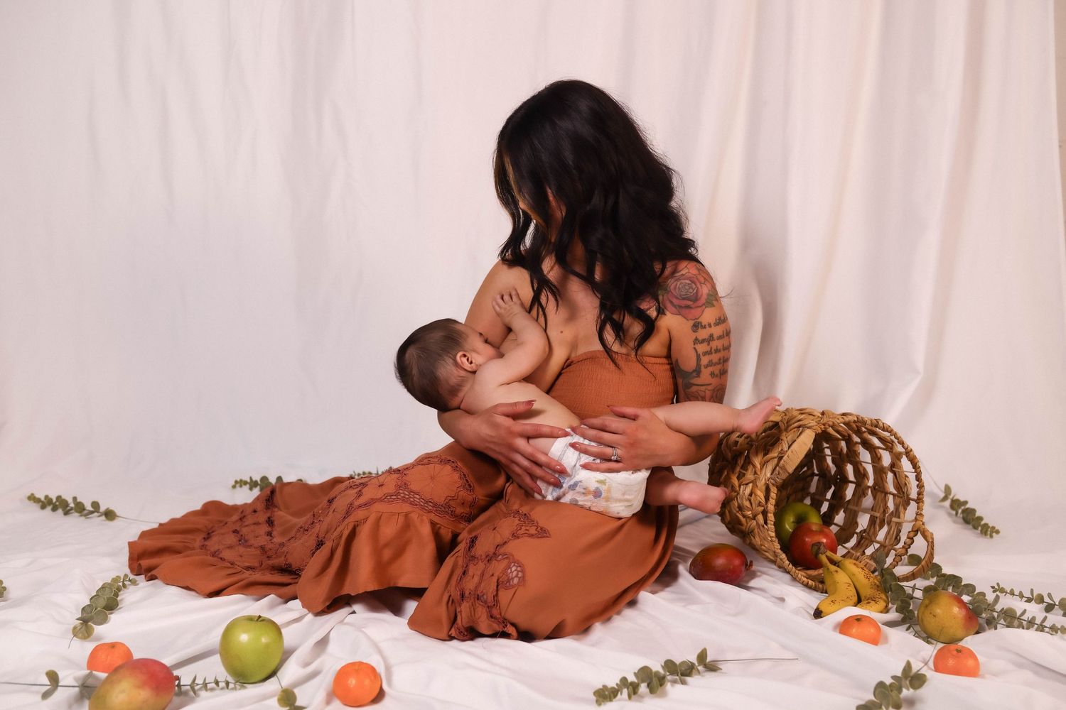 A mom and baby breastfeeding on a white backdrop with scattered fruit and flowers around them, wearing matching rust-colored outfits.