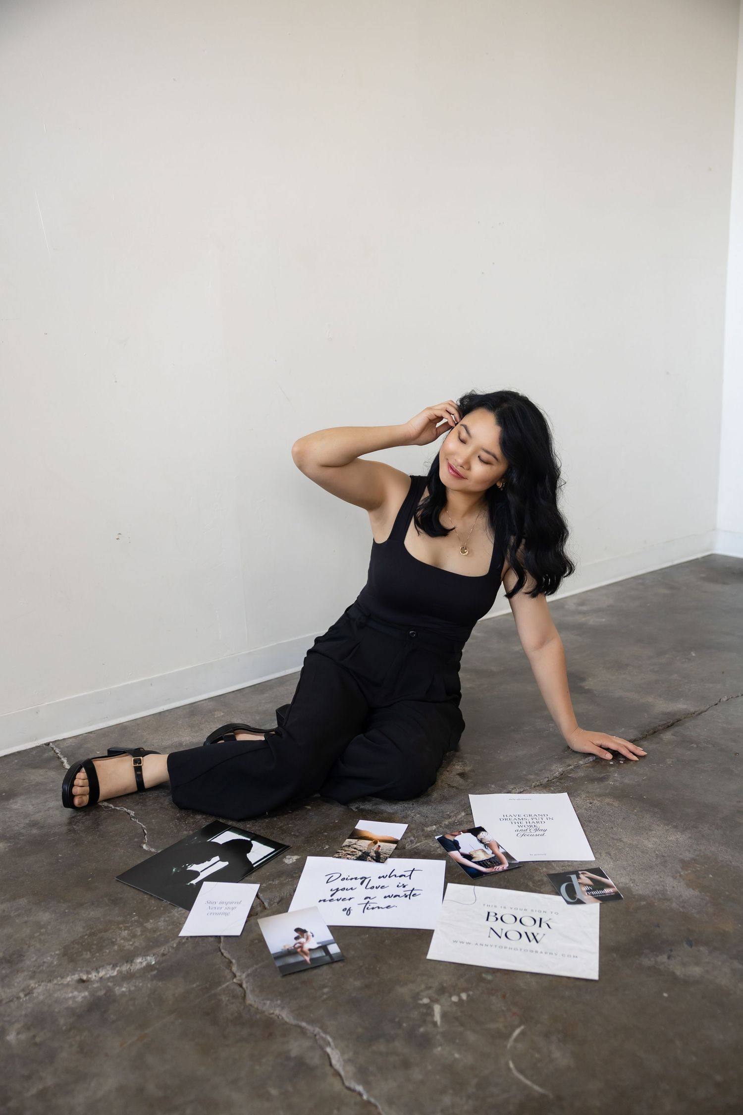 Person in black business outfit sitting on concrete floor with scattered photos and quotes in artistic pose while holding hair back.