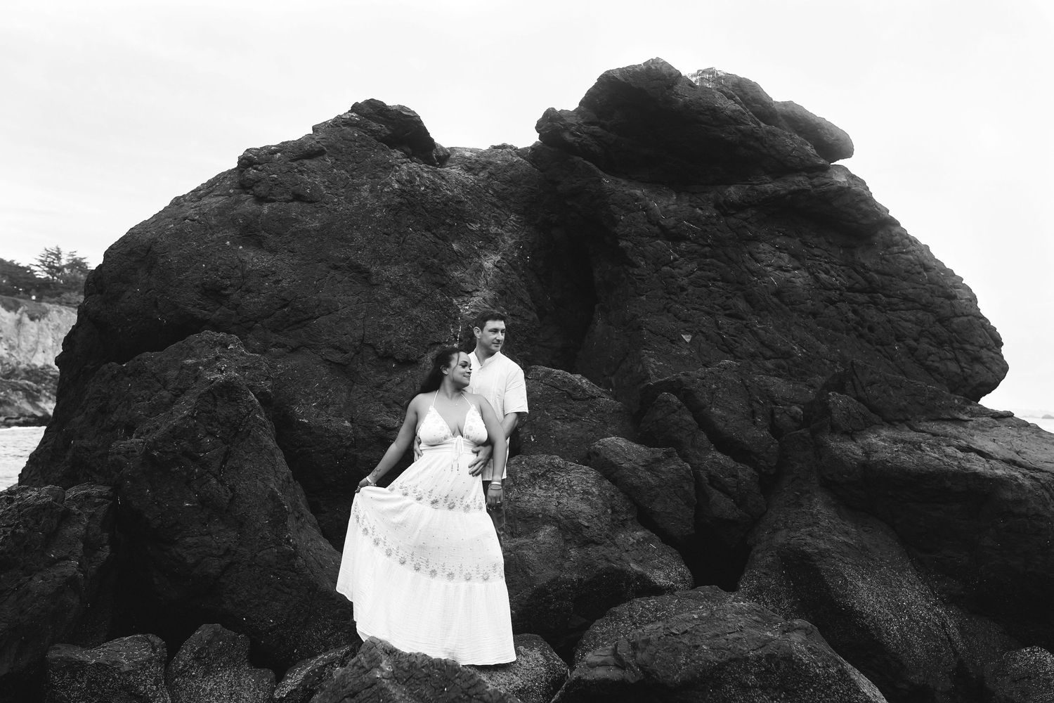 A series of couples dressed in white attire pose on dramatic black lava rocks near the ocean at sunset.