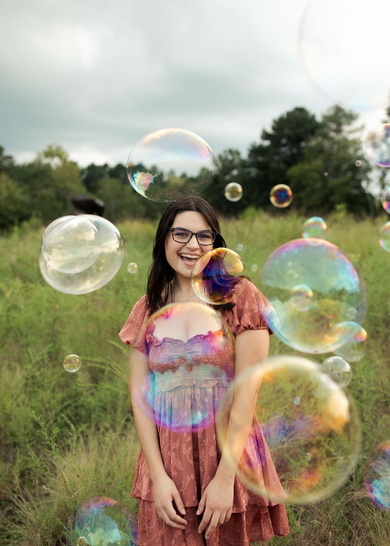 A girl is photographed surrounded by colorful soap bubbles floating through the air in a grassy field on a sunny summer day.